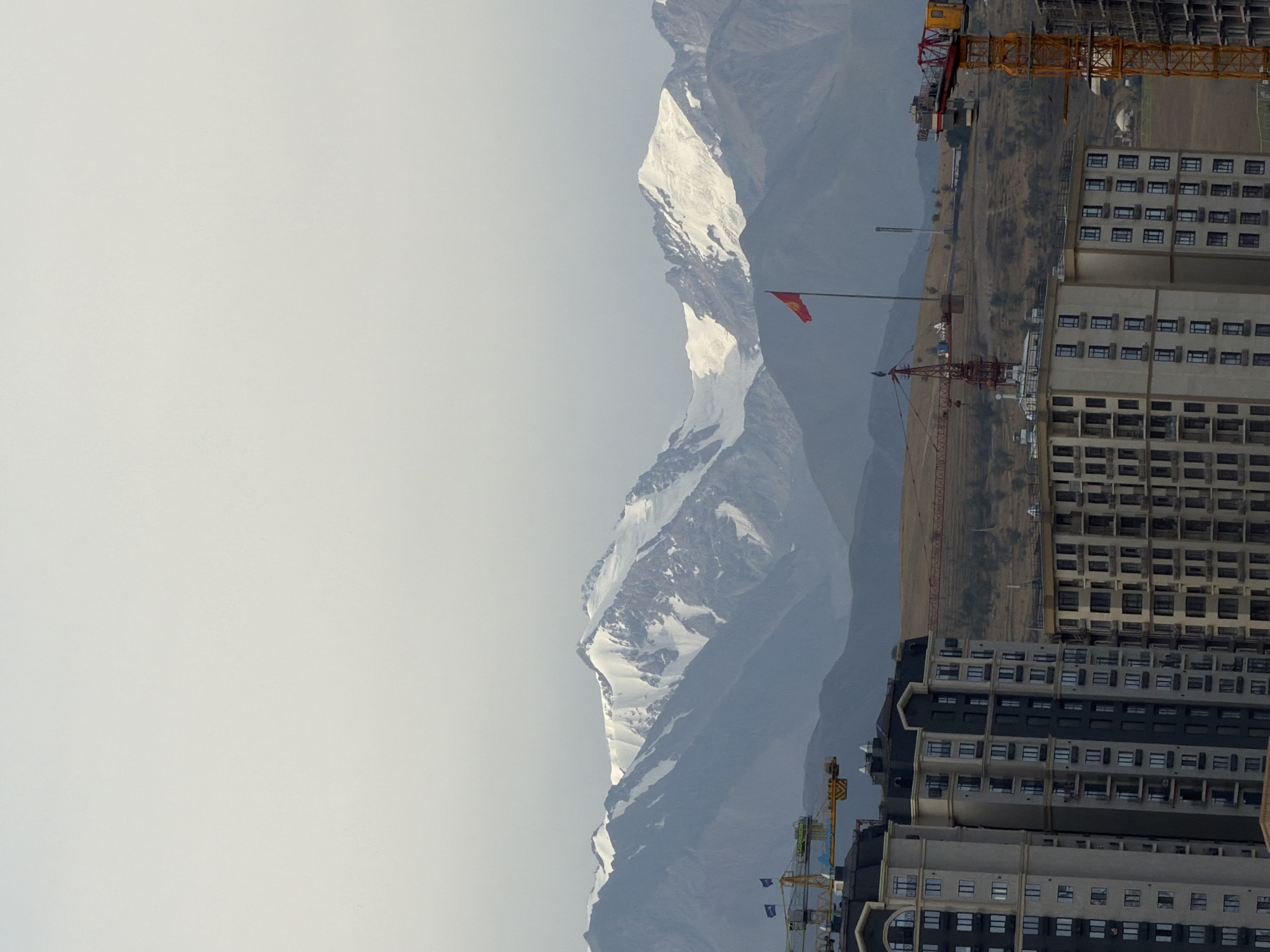 The image captures a cityscape against a backdrop of majestic, snow-capped mountains under a hazy sky. In the foreground, several modern, multi-story apartment buildings are under construction, with their facades showing a grid of windows and balconies. Construction cranes, some yellow and some red, are visible among the buildings and on the elevated, dry, hilly terrain behind them. A red flag, possibly a national flag, flies from a tall pole on the hill. The scene suggests an urban development project in progress, juxtaposed with the natural grandeur of the mountains.