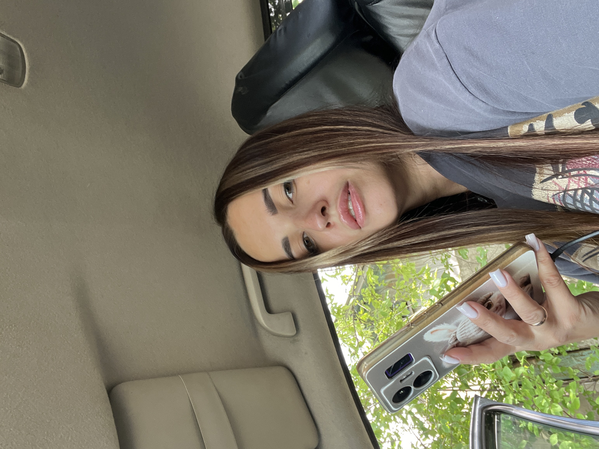 In the interior of a vehicle, a woman with long, dark hair with blonde highlights is holding a smartphone in her left hand. She is looking at the phone with a neutral expression. The woman is wearing a grey t-shirt. The interior of the car is visible, including the headliner and a portion of the passenger seat. Outside the car, green foliage is visible through the window.