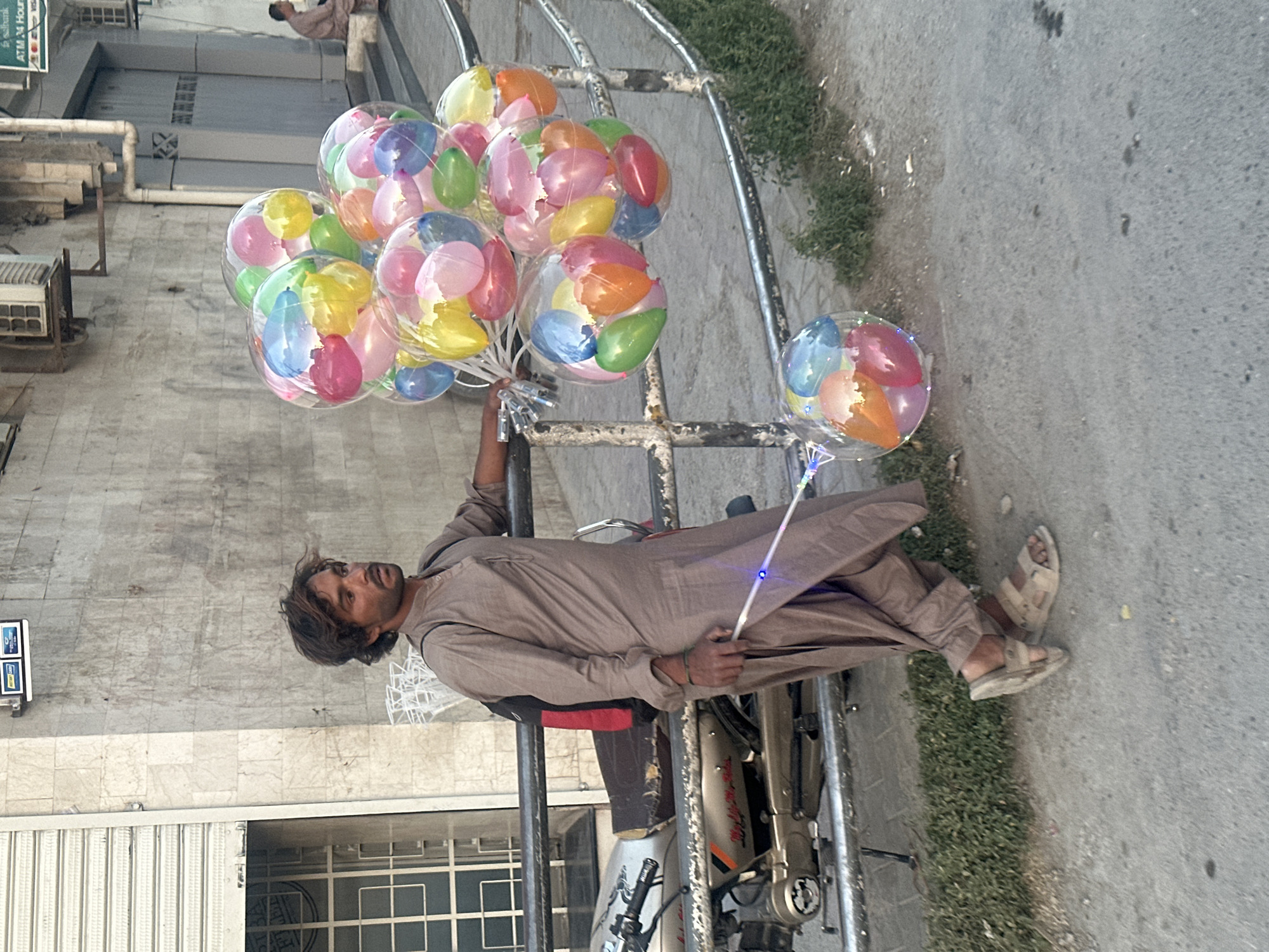 A man in Quetta City Tehsil, Pakistan, wearing a light brown shalwar kameez and sandals, sits on a parked motorcycle holding a large bunch of colorful balloons inside clear plastic. The motorcycle is positioned on the sidewalk near a building with a closed metal-grille door, a partially visible window, and an air conditioning unit. A strip of grass separates the sidewalk from the asphalt road.