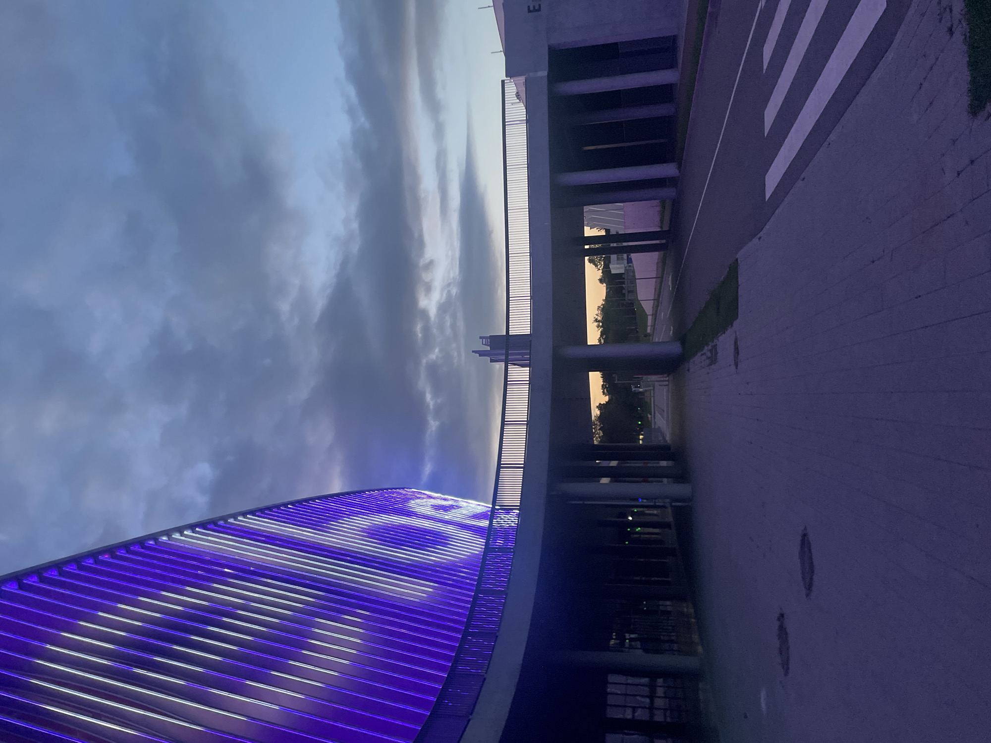 This is an eye-level, wide shot of a modern building facade illuminated with purple and white lights, curving into the frame from the left. The building's exterior features vertical slats, many of which glow with light. In the midground, a walkway with a metal railing bridges over a road. The walkway is supported by large, cylindrical columns. The road features a white pedestrian crossing. The sky above is cloudy and appears to be twilight, with hints of orange near the horizon. The ground in the foreground is paved with interlocking stones. The scene appears to be an exterior view of an entertainment venue or stadium.