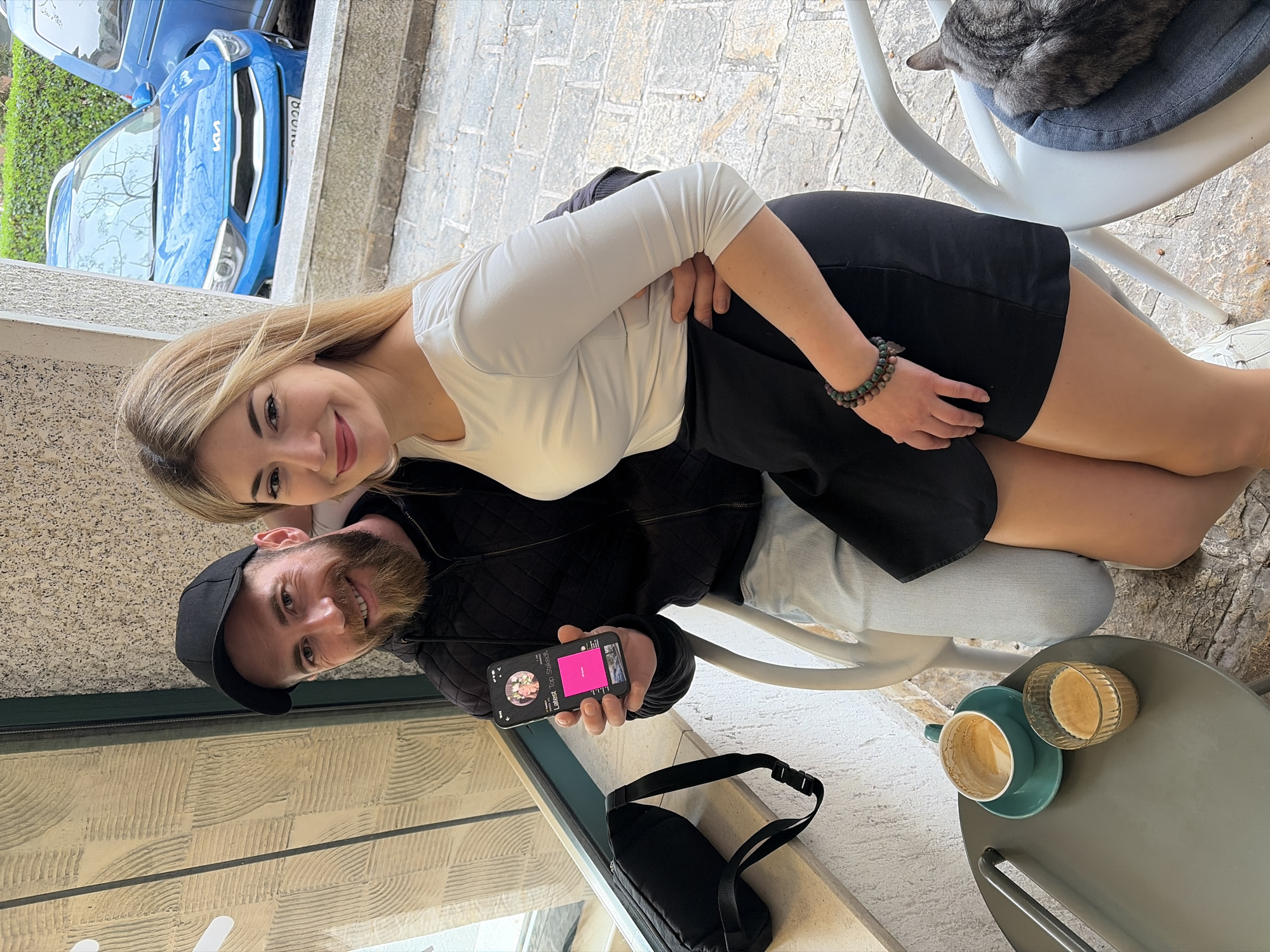 The photograph captures a man and a woman posing closely together at an outdoor café in Budva, Montenegro. The woman, with blonde hair and a bright smile, is wearing a white long-sleeved shirt and a short black skirt, with a beaded bracelet on her wrist. The man, sporting a beard and a black baseball cap, is holding a smartphone with a screen displaying "Latest for Sehnsucht" alongside floral imagery and a pink progress bar.
They are seated near a gray table where a turquoise coffee cup with some remaining liquid and a glass container are present. The man is wearing a black quilted jacket and light-colored jeans. A black bag with a strap is visible near the table.
In the background, a blue Kia car is partially visible next to a wall covered in green foliage. A cat is curled up on a chair cushion. The café's surroundings include textured walls, likely reflecting the local architecture, and a stone-paved surface. The café is located near a window.