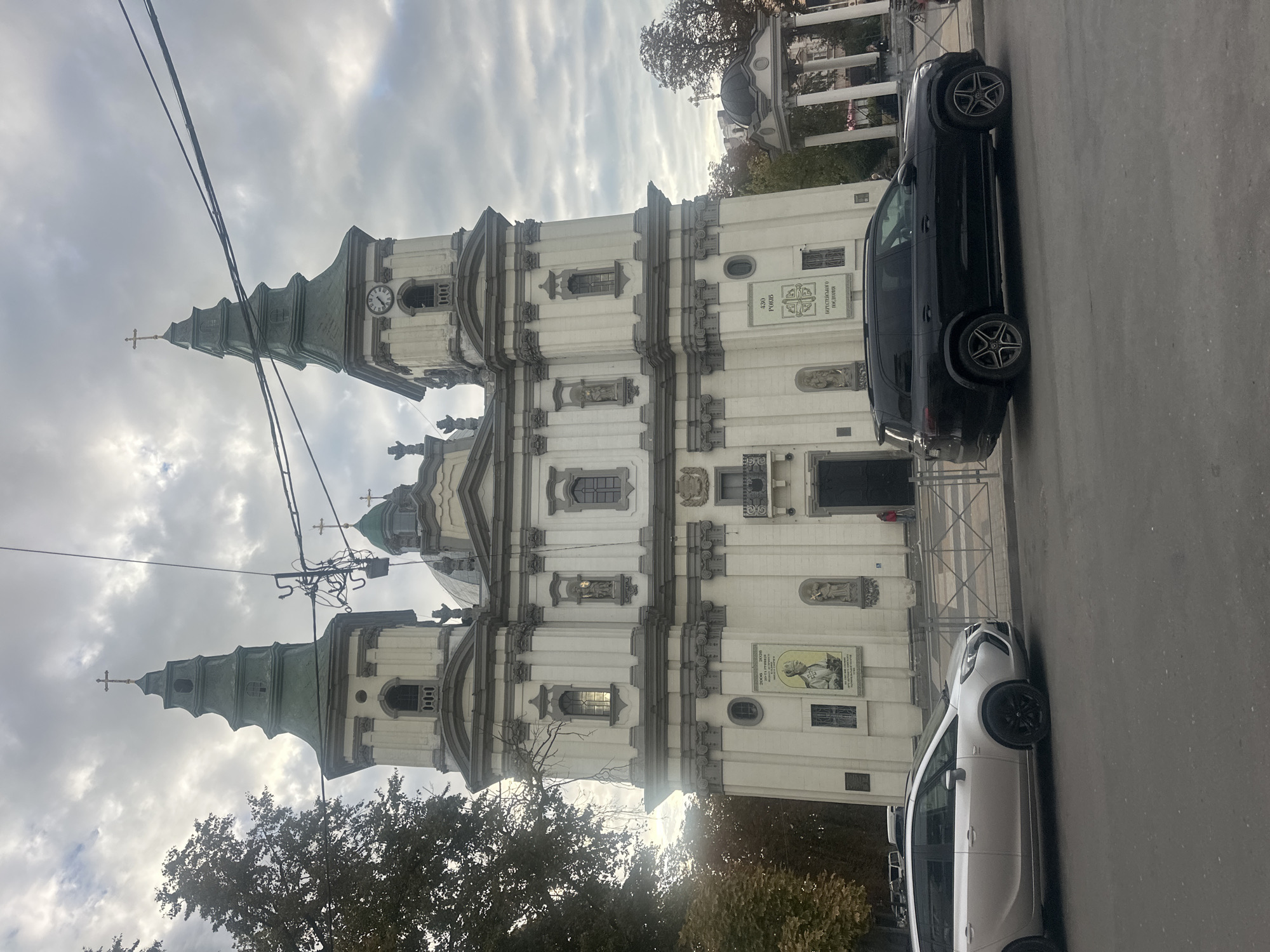 The image shows the Church of the Immaculate Conception of the Blessed Virgin Mary in Ternopil, Ukraine, on a cloudy day. A silver station wagon is parked in front of the church to the left, and a black SUV is parked to the right. The church features two prominent towers with green spires, one of which has a clock. The facade is white with gray architectural details and several statues and decorative elements. There are signs on the church walls, including one with a portrait of a religious figure. The sky is overcast with dramatic clouds, and trees with autumn foliage are visible on the left side of the image. Overhead power lines are also present in the scene.