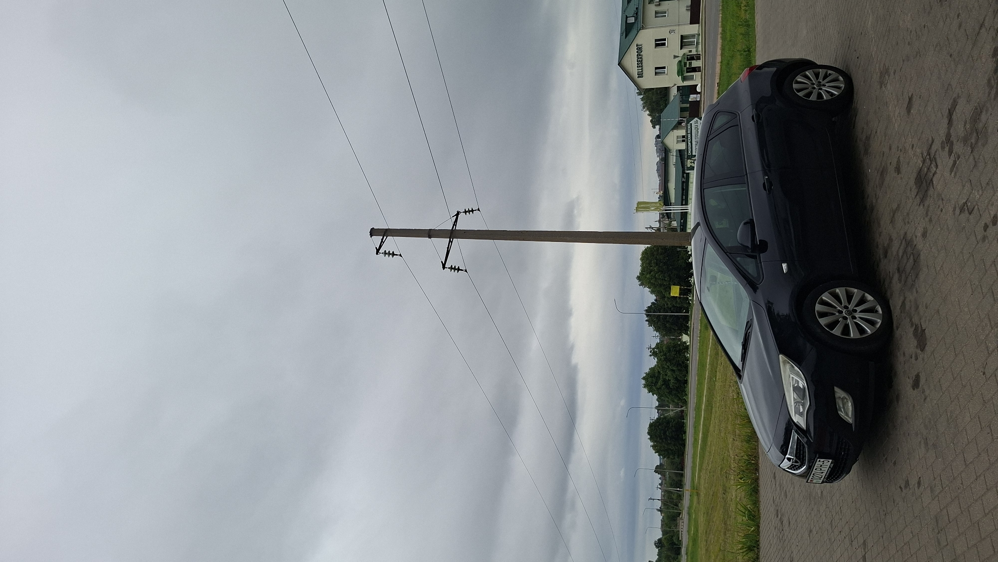 A dark blue Opel Astra hatchback is parked on a paved surface in front of a building with a green roof and the sign "BELLESEXPORT" in Смалявічы, Belarus. The car is positioned diagonally, facing left. In the background, there is a tall concrete utility pole with electrical wires extending from it. The sky is overcast with dark clouds, and there are trees and streetlights visible along a road in the middle distance. The overall scene depicts a roadside parking area on a cloudy day.
