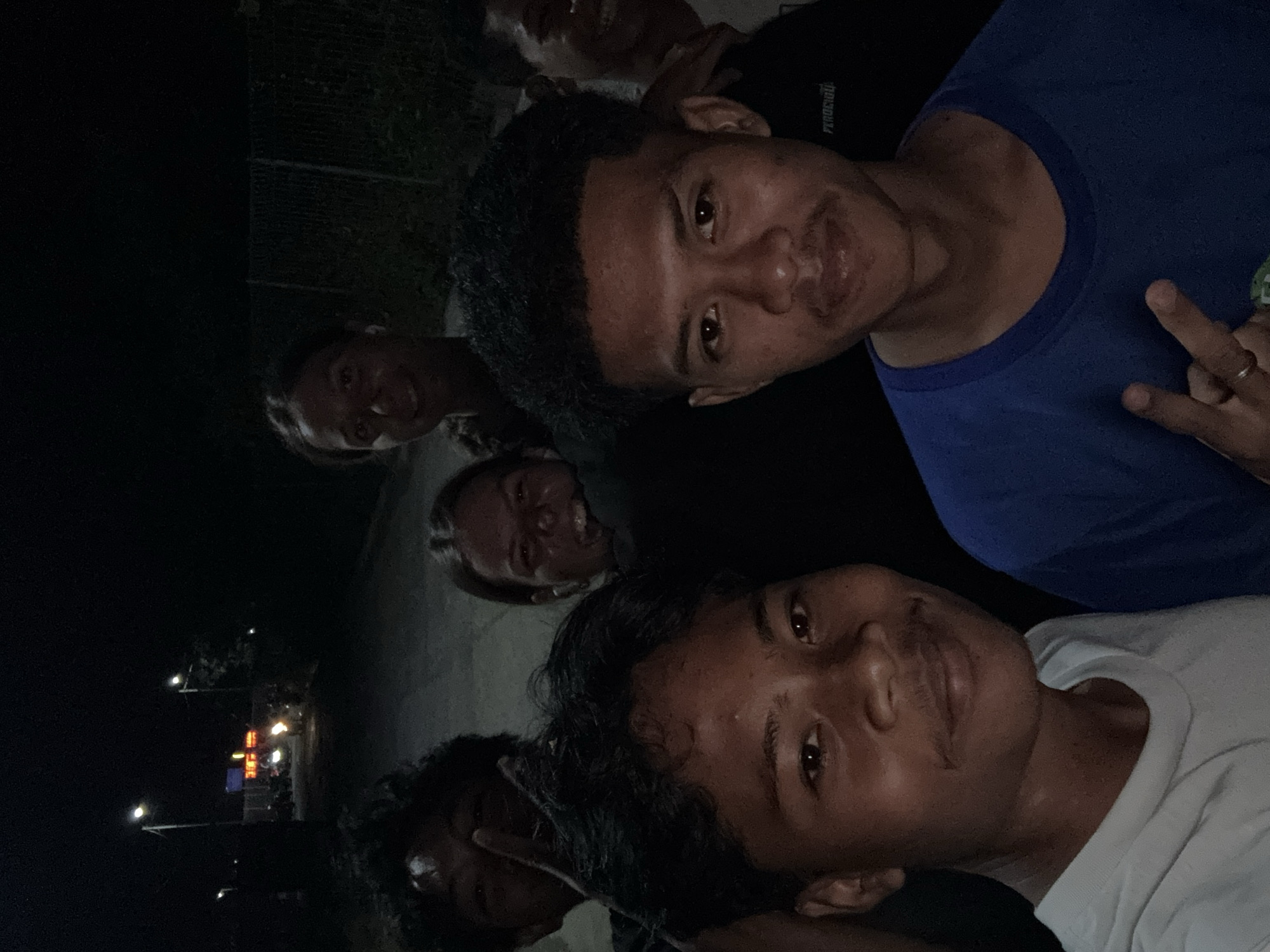 The image depicts a group of young people outdoors at night, possibly in Labuhan Lombok, Indonesia, given the context. Several individuals are visible, with varying degrees of focus. The lighting suggests a street or open area, with a few light sources casting illumination. In the background, a street with a few vehicles and lights can be seen, along with what appears to be a fence. Some of the individuals are smiling and looking towards the camera, while others are partially obscured. The overall impression is a candid snapshot of friends gathered at night.