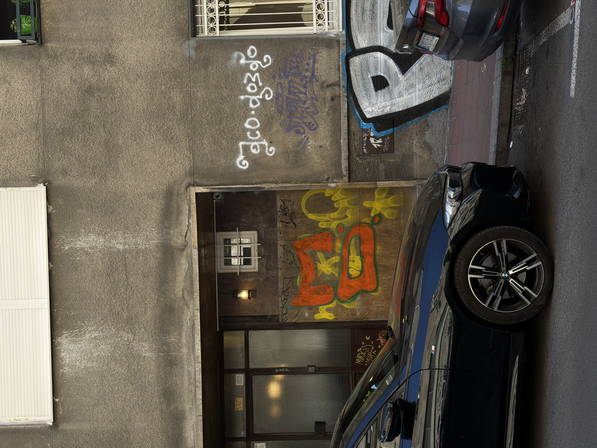 The image depicts a narrow alleyway in Belgrade, Serbia. The alleyway is flanked by two buildings, both covered in graffiti. One building has a white shuttered window and a doorway that leads into a dark interior. Above the doorway is a small window with a barred frame. Graffiti on the walls includes lettering in various colors and styles. Two cars are parked on the right side of the alleyway, partially blocking the view. The pavement is a mix of brick and asphalt.