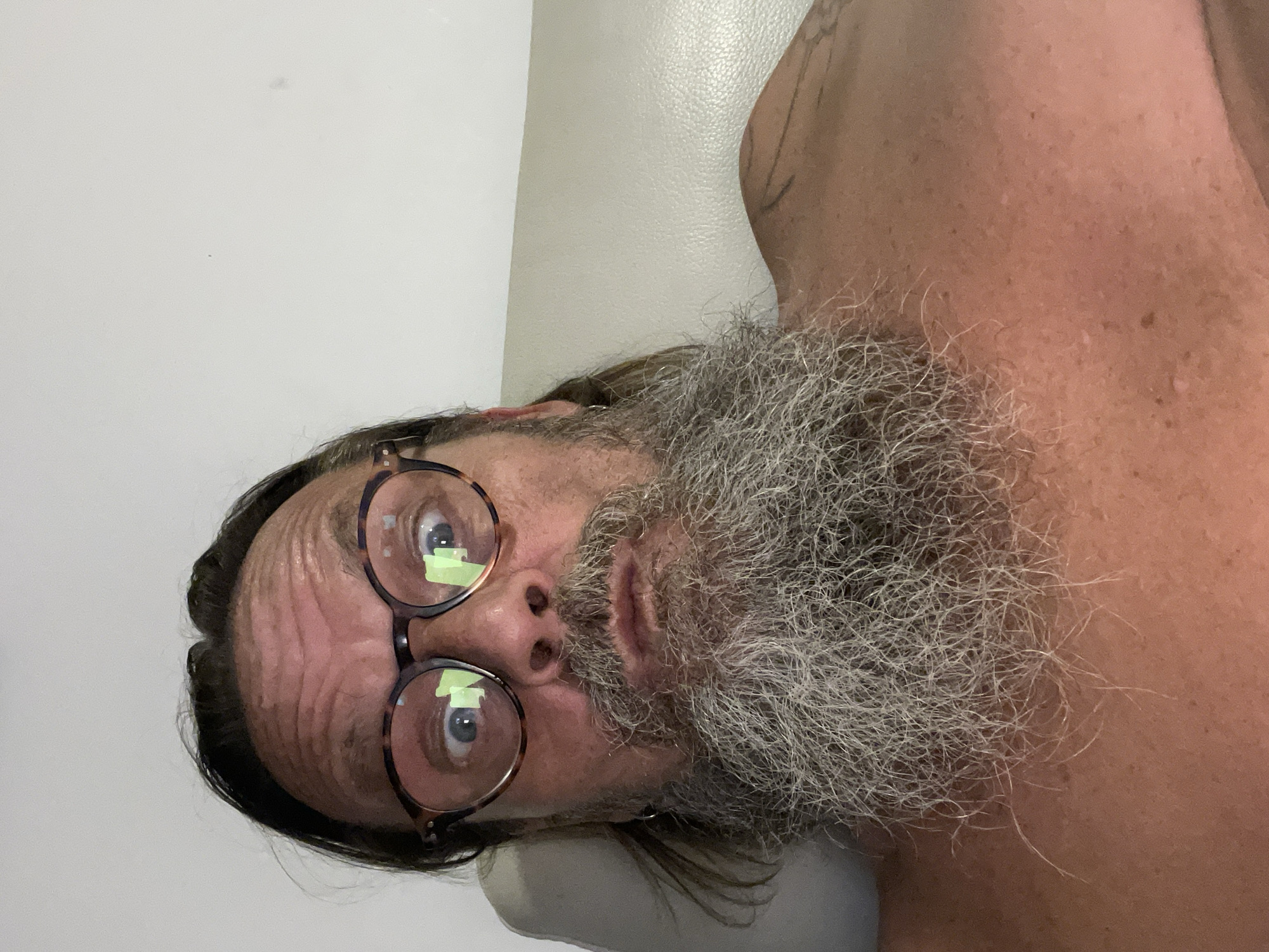 A close-up, selfie-style photograph shows a man with long, dark hair, a grey beard, and tortoiseshell glasses. His eyes are wide, and his mouth is slightly open, giving him a surprised or bewildered expression. The man is shirtless, revealing a tattoo on his right shoulder and upper arm. He appears to be reclining on a light-colored, possibly leather, surface. The background is a plain, light-colored wall. The lighting suggests the photo was taken indoors, possibly at night, with a screen's reflection visible in his glasses. No specific location in Netanya, Israel, can be identified from the image.