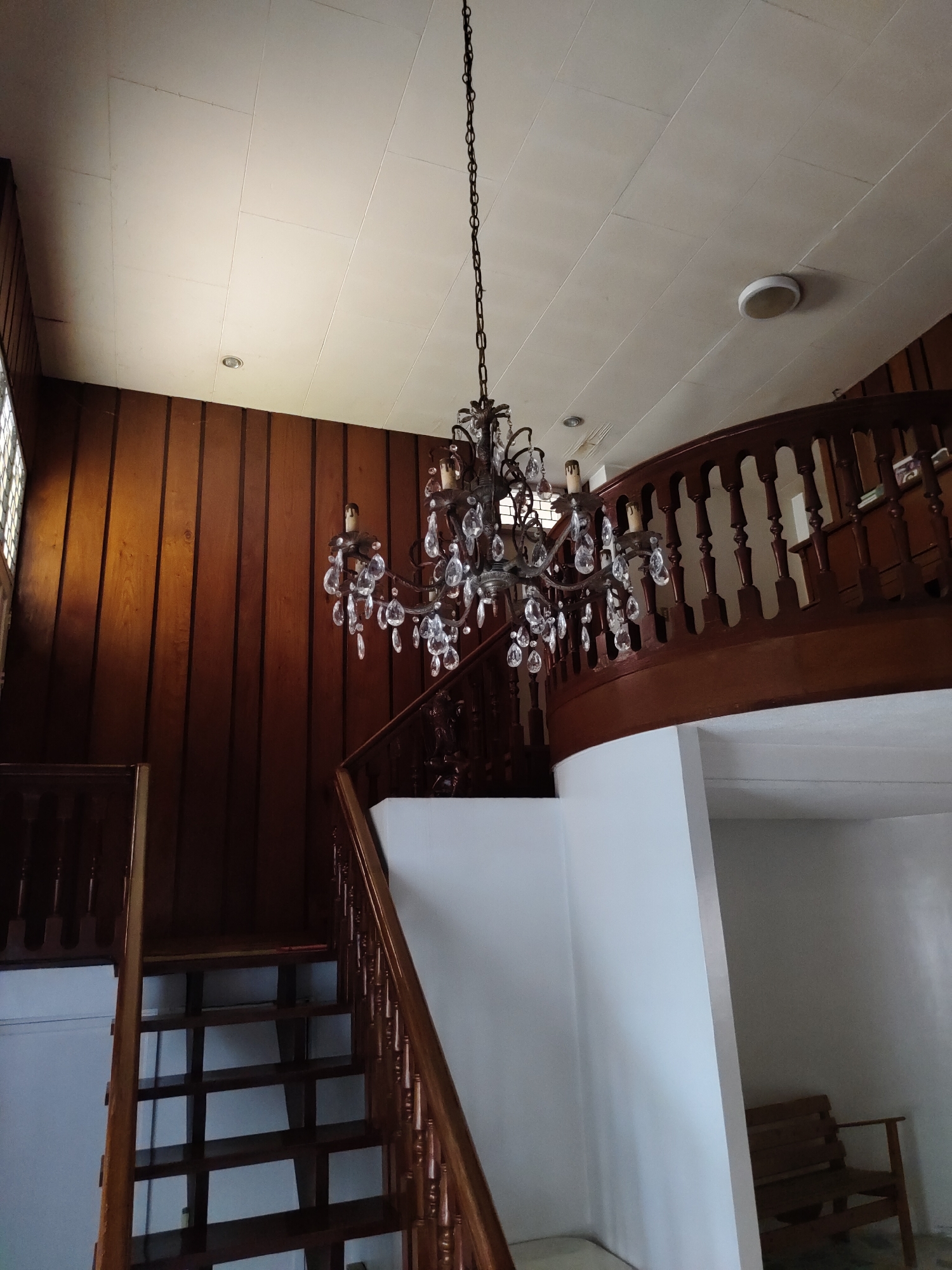 An interior shot depicts a multi-level building in Davao City, Philippines, featuring a grand wooden staircase and a prominent chandelier. The scene is dominated by a dark wood-paneled wall on the left and center, contrasted with a white, paneled ceiling. A large, multi-armed chandelier, adorned with numerous clear crystal drops and candle-style lights, hangs centrally from the ceiling by a long chain. The staircase, constructed of dark wood, includes a grand, curved balustrade with turned balusters and a handrail extending upwards and curving to the right. A lower section of the wooden staircase is visible on the left, displaying multiple steps. The white ceiling contains at least two circular light fixtures, one recessed and one surface-mounted, with visible panel seams showing minor discoloration. A grid-patterned window is partially visible on the far left. On the lower right, a dark wooden bench is partially visible within a recess, and indistinct objects are visible on a ledge behind the upper balustrade. No individuals are present in the image.