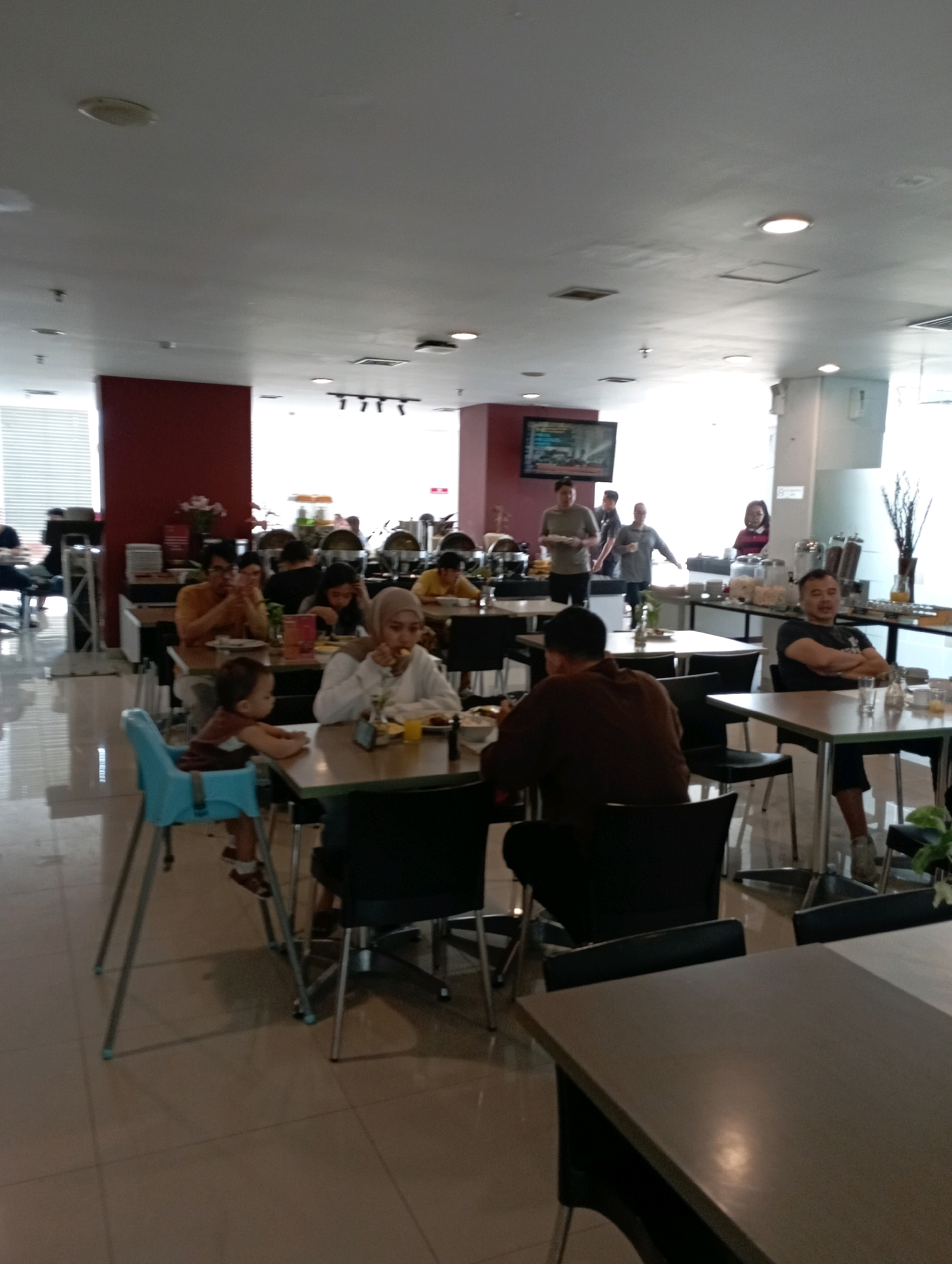 The image depicts an indoor dining area, likely a hotel breakfast buffet, possibly located in Malang, Indonesia. Several tables are occupied by individuals and families. In the foreground, a child is seated in a blue high chair at a table. To the right, a man in a dark shirt is seated at a table, looking towards the camera. Further back, several groups are seated at tables, some eating, others conversing. A buffet line with various food service equipment is visible in the background, with staff attending to it. The environment appears to be well-lit with overhead lighting.