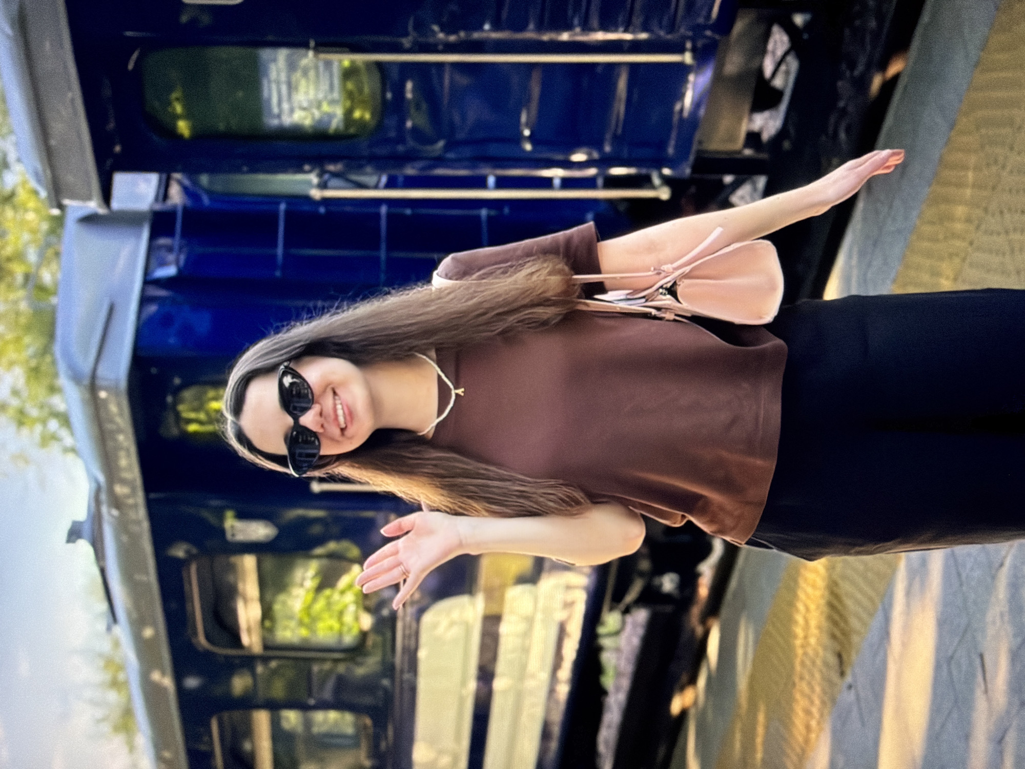 A young woman wearing sunglasses, a brown t-shirt, and black pants stands on a train platform, smiling and waving. She has a small pink purse slung over her shoulder. Behind her, a blue train is visible, with green trees and a blue sky in the background. The platform has yellow tactile paving.