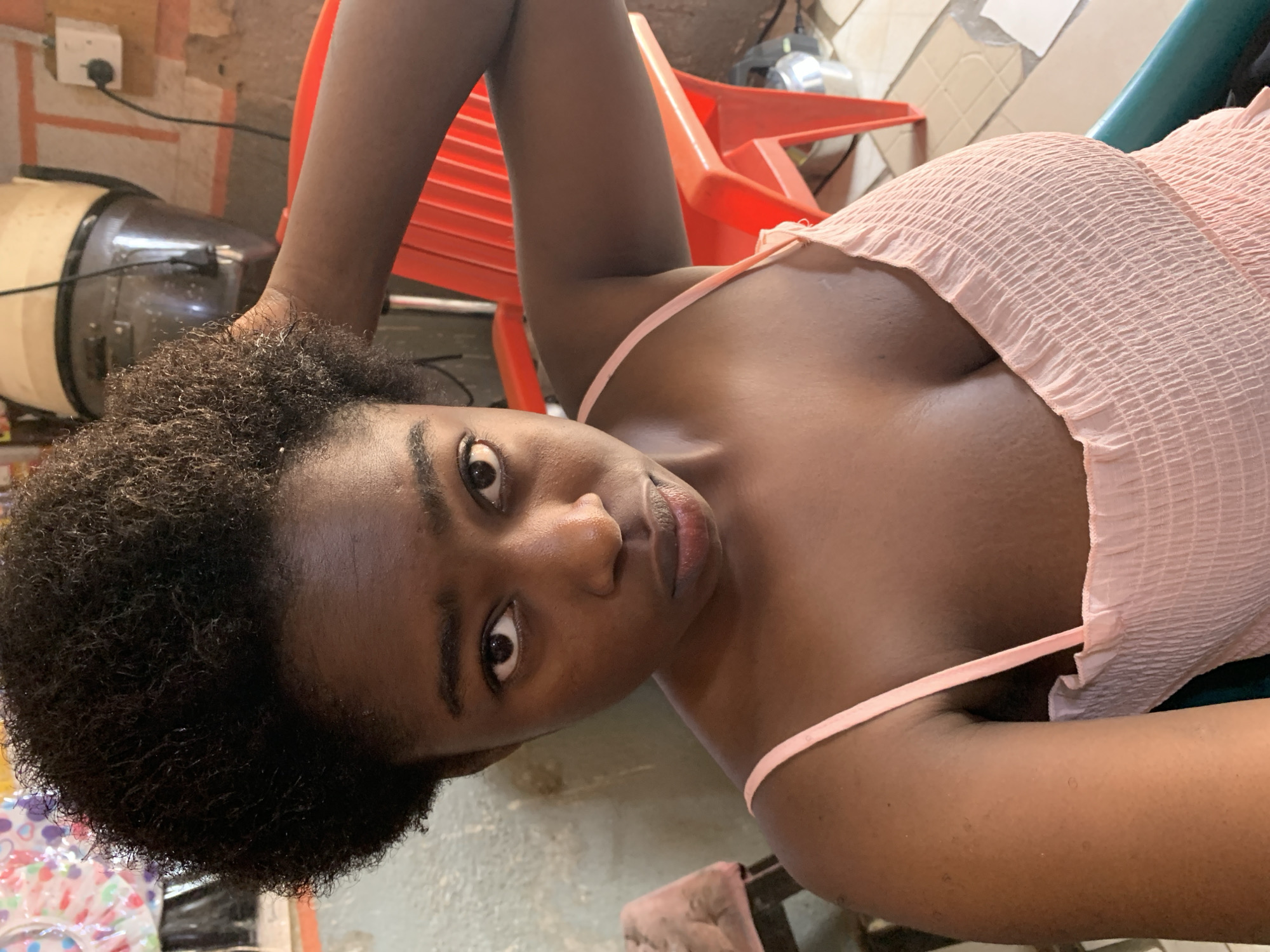 A dark-skinned woman is seen close-up, lying on a light-colored tile floor. She has tightly curled dark hair and is wearing a light pink, smocked top with thin straps. Her head is turned towards the camera, and she appears to be looking directly at the viewer. In the background, there is a red plastic chair, a hair dryer and a wall with exposed brickwork.