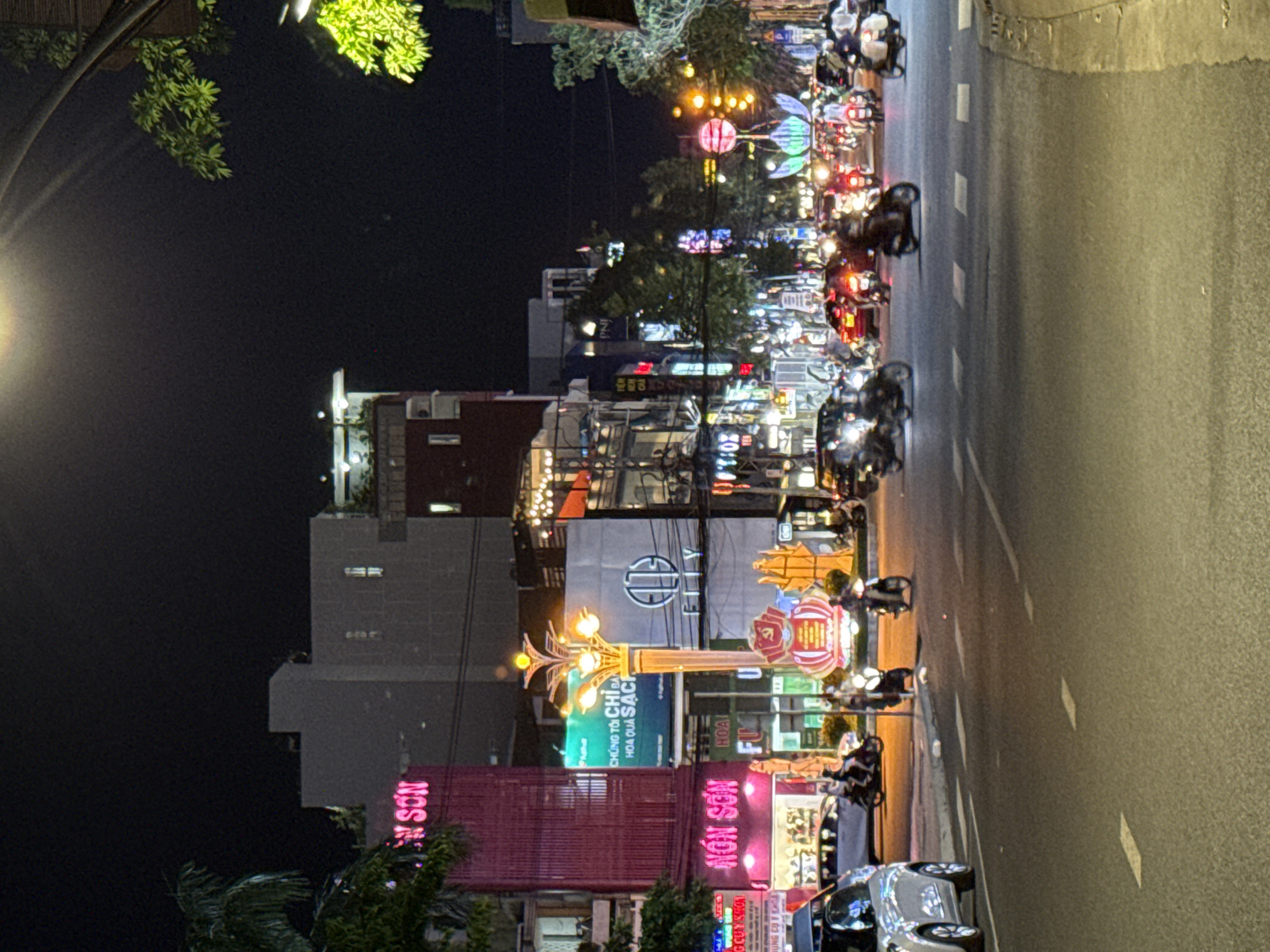 This is a nighttime photograph of a busy street in Nha Trang Ward, Vietnam. The scene is illuminated by the bright neon signs of various businesses and the headlights of passing vehicles. The street is filled with motorcycles, suggesting a common mode of transportation in the area.
The buildings lining the street display a vibrant mix of advertisements and signage, with a prominent pink neon sign that reads "NOS SON" in Vietnamese. Another sign, also in Vietnamese, translates to "We only sell clean fruits." The overall atmosphere is lively and bustling, typical of a commercial street at night. The lighting creates a festive and somewhat chaotic ambiance.
The time of day is clearly night, as indicated by the dark sky and the artificial illumination from signs and vehicle lights. The weather appears to be clear, as there are no visible signs of rain or inclement conditions. The presence of multiple people on motorcycles indicates ongoing activity and movement on the street. The photograph captures a slice of urban life in Vietnam, highlighting the energetic street scene and commercial activity.