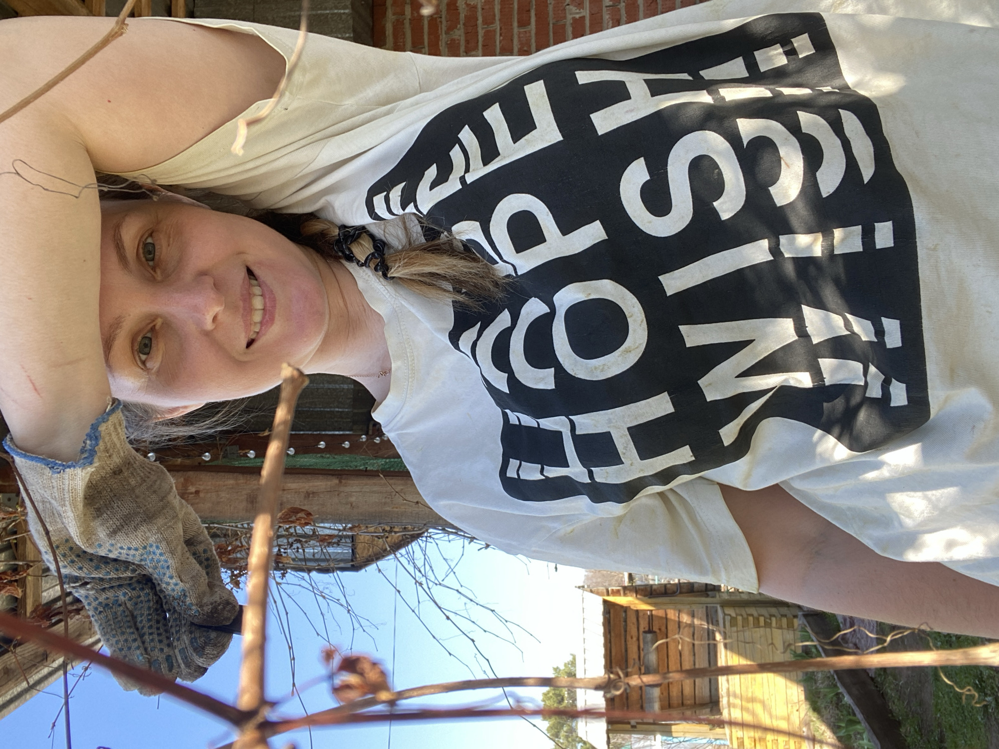 A fair-skinned woman wearing a light-colored t-shirt with bold black lettering is outside, pruning branches with gloved hands in Chaikovsky, Russia. The background includes brick and wooden structures, as well as clear blue sky visible through the branches.