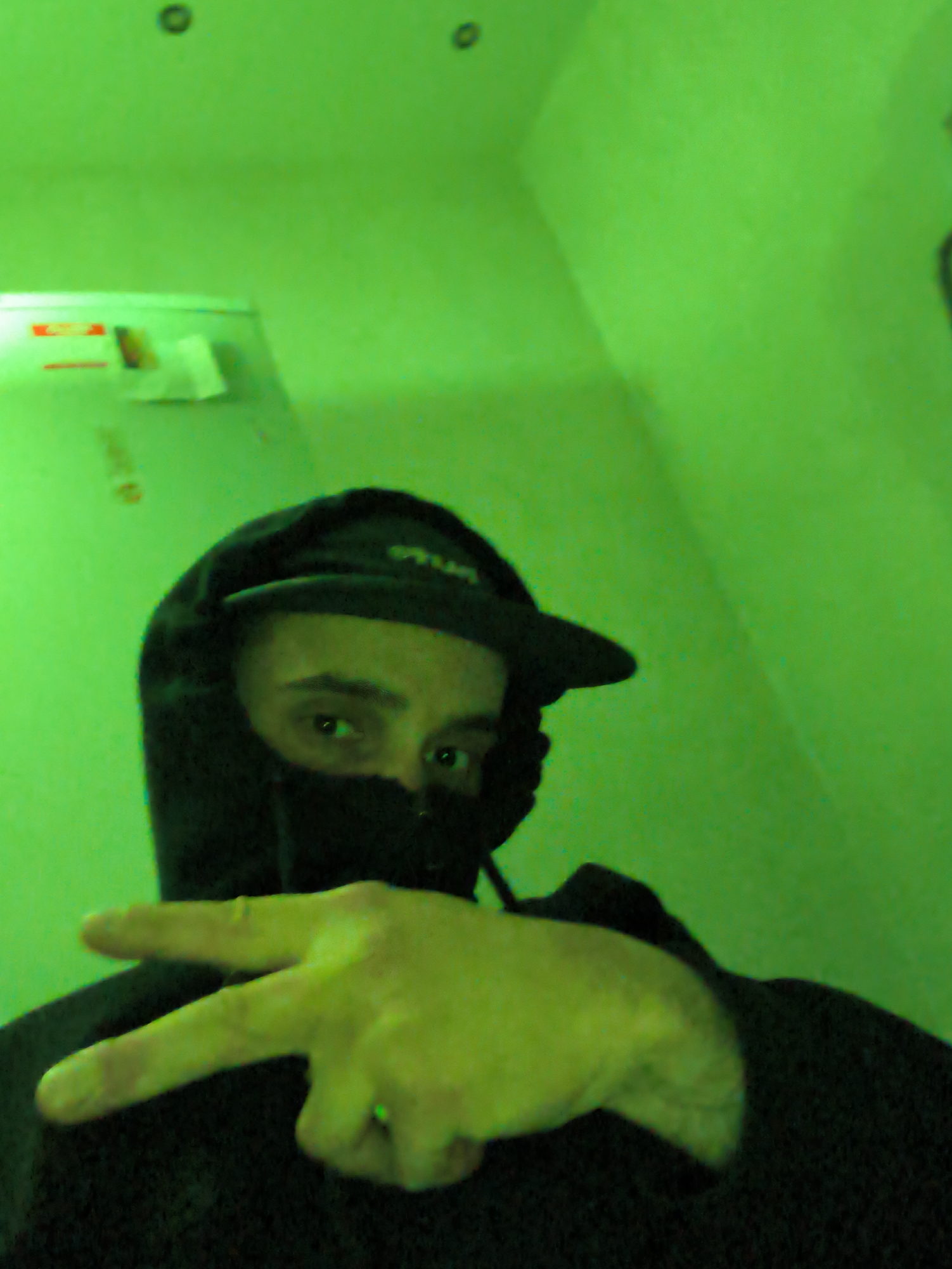A person wearing a black hooded sweatshirt and a black face mask is posing for a selfie. They are holding up their hand, making a "peace" sign. The background is a green wall. The location, Hrodna, Belarus, cannot be determined from this image.