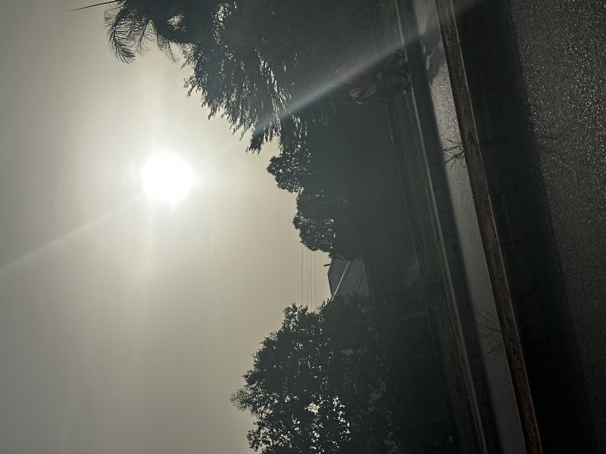 The image shows a view from what appears to be a vehicle, likely a car, in Bukuru, Nigeria. The sky is bright and overcast, with the sun visible as a glaring white disc. Trees line the side of the road, casting shadows. A building is also visible behind the trees. To the right of the image is the vehicle window and frame. The quality of the image is poor.