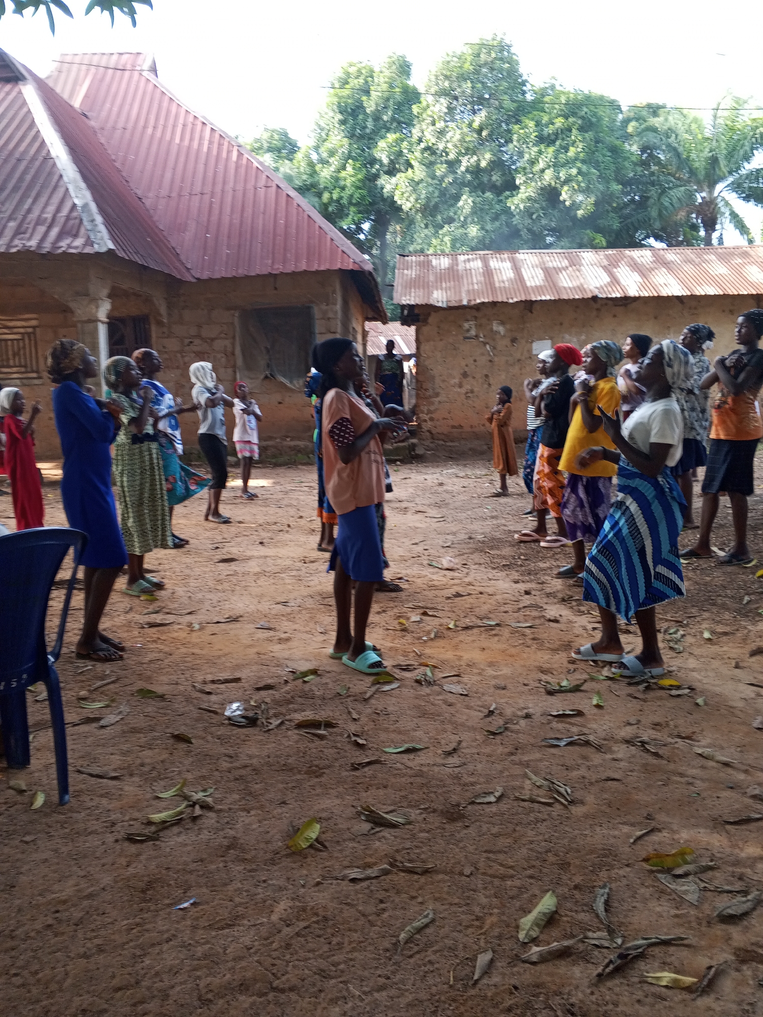 A group of about 20 people, mostly women, stand in a dirt yard in Akwanga, Nigeria. Two buildings with corrugated metal roofs are visible behind them. Many of the women are wearing headscarves and colorful skirts or dresses. They appear to be looking in the same direction, possibly listening to someone who is out of the frame. Leaves are scattered on the ground. A blue plastic chair is visible on the left side of the frame.