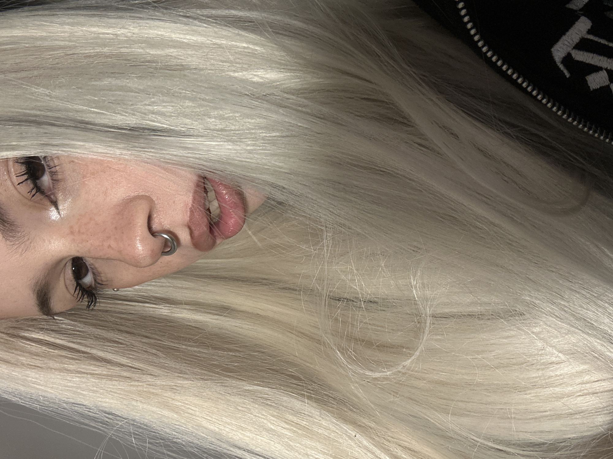This medium close-up portrait, taken in Buenos Aires, Argentina, features a person with striking, very long, platinum blonde hair that dominates the frame. The subject's head is tilted, with the photo taken from an overhead perspective as if the person is looking up at the camera while lying down.
Key facial features visible include one dark brown eye with long, dark, mascara-laden eyelashes and black eyeliner. Freckles are scattered across the nose and cheek area. A silver septum ring adorns the nose, and a small silver stud piercing is visible below the eye. The lips are slightly parted, revealing a glimpse of teeth and appearing to have a subtle gloss.
The extensive, straight, and lustrous blonde hair fills most of the image, creating a strong visual impact. In the upper right corner, a segment of a black garment is visible, featuring a zipper with small silver teeth and stylized white embroidery that is partially obscured and indistinct.
The lighting is soft and even, highlighting the texture of the hair and the subtle details of the skin. The overall mood is cool and stylish, with the focus strongly on the subject's distinctive features and hair.