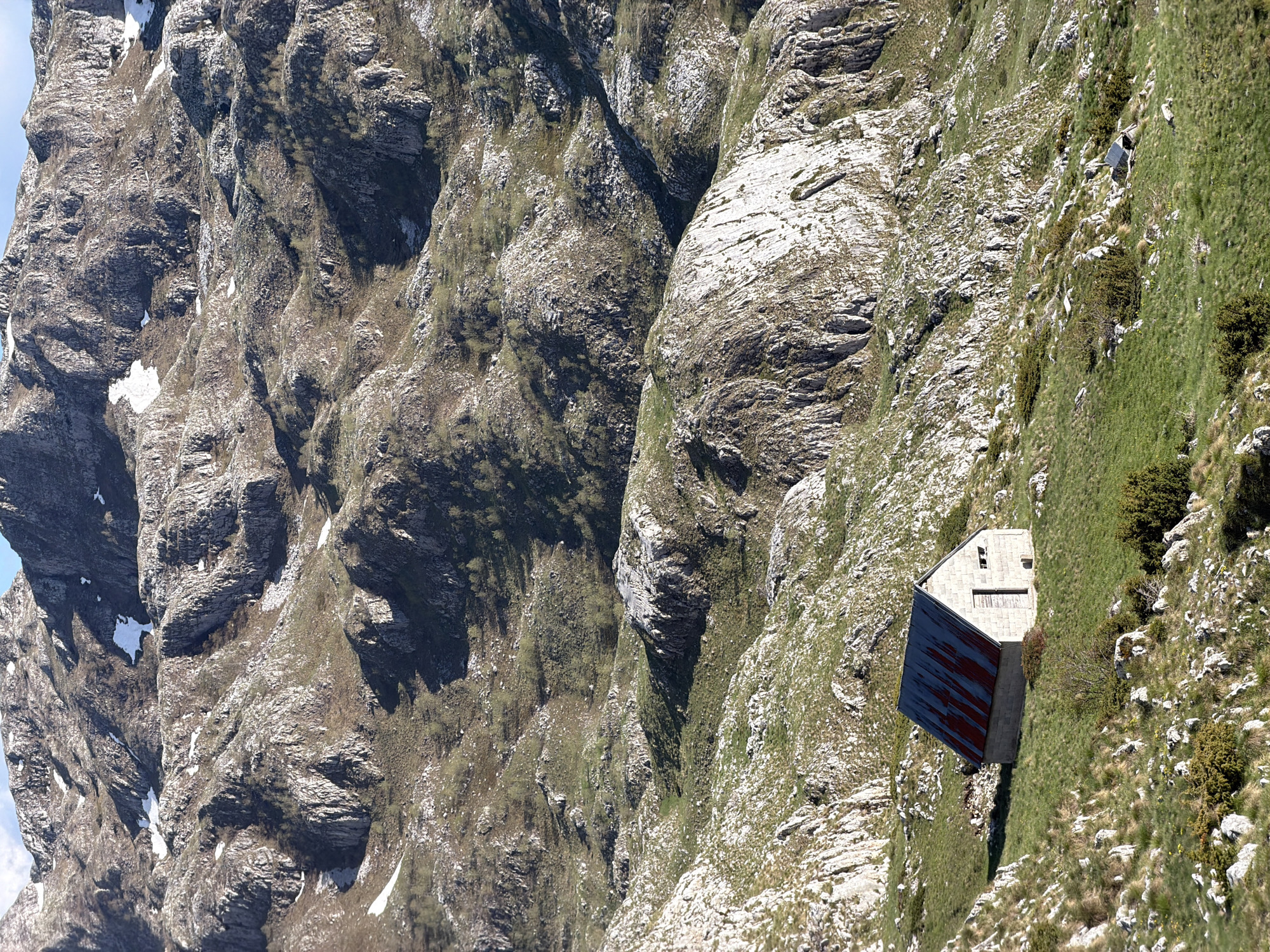 A mountain scene with a small building. The building is perched on a steep, grassy slope, with rugged, rocky mountains on either side. The building has a grey stone structure with a dark red or blue roof. The area around the building is covered in green grass and small shrubs, contrasting with the bare rock. The mountains are rugged and rocky, with patches of green moss and some snow. The image gives a sense of remoteness and harsh environment. Location is Boričje, Montenegro.