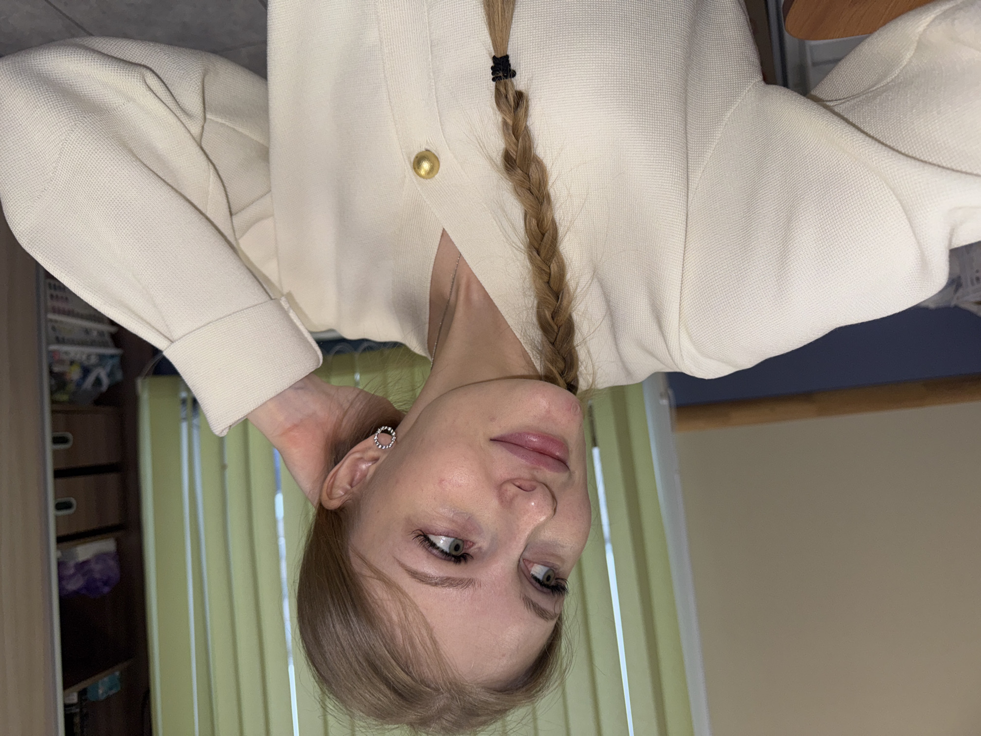 A young woman is centered in the frame, positioned at an inverted angle, with her head nearly touching the bottom edge of the image. She has light-colored hair styled in a braid and light skin. She is wearing a light-colored cardigan with a gold button. Her left hand is positioned near her ear.
The background consists of light green vertical blinds and a light tan wall. A wooden desk is visible in the upper right corner. A storage unit with drawers is present in the left background, with items stored inside. The photograph appears to be taken indoors.
The photograph was not taken in Perm, Russia, as there is no evidence of any Russian city.