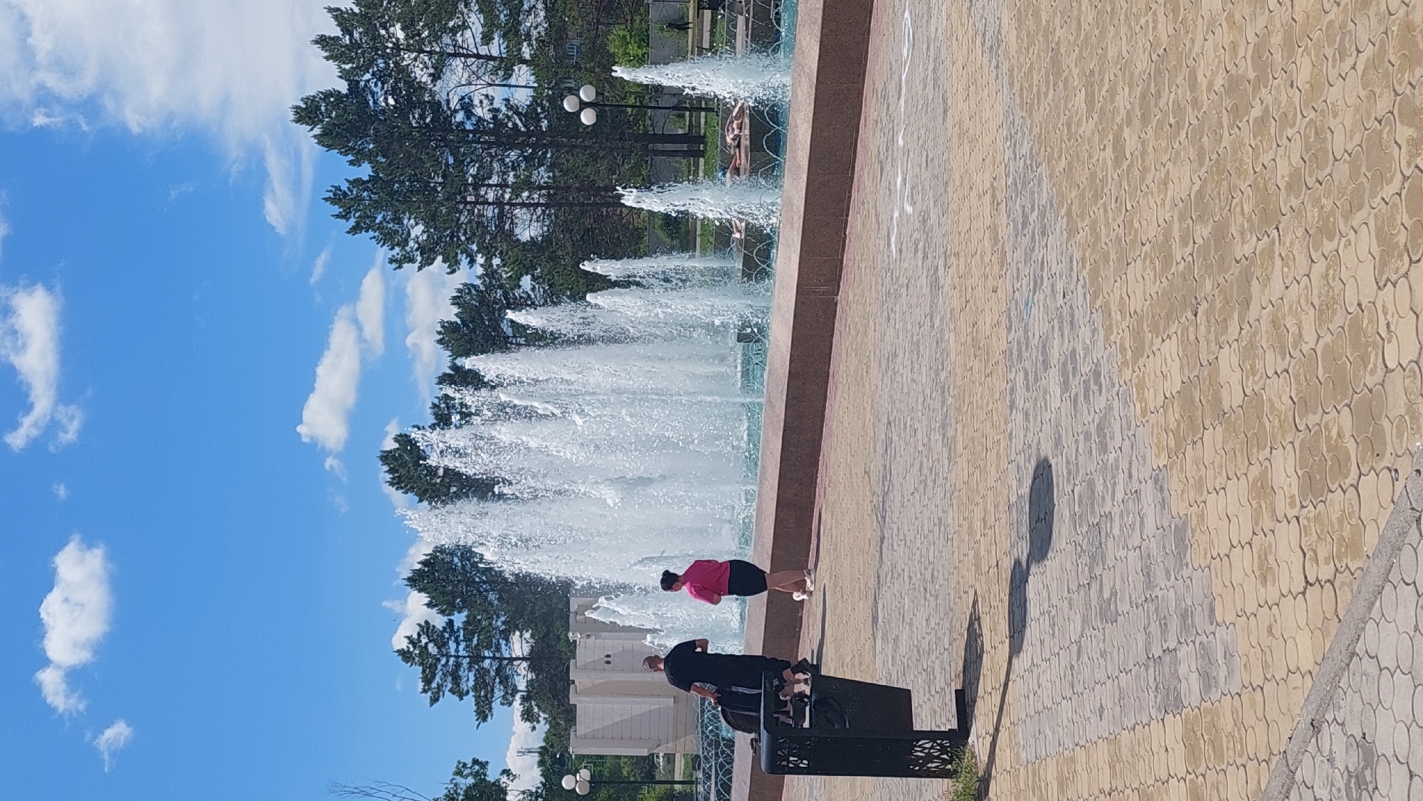 The image depicts a large, multi-tiered fountain in a paved public area, likely a park or plaza. The fountain is active, with numerous jets of water creating a dynamic display. In the foreground, a black trash receptacle is visible on the left. Two individuals are walking away from the camera, towards the fountain. One, wearing a black t-shirt and dark pants, is pulling a black bag on wheels. The other, wearing a pink top and dark shorts, walks alongside. The pavement is made of interlocking tan and gray pavers. In the background, a line of coniferous trees forms a backdrop, with a light-colored building and streetlights visible behind them. The sky is blue with scattered white clouds. The scene appears to be in Komsomolsk-on-Amur, Russia, based on the common architectural and landscape elements observed.