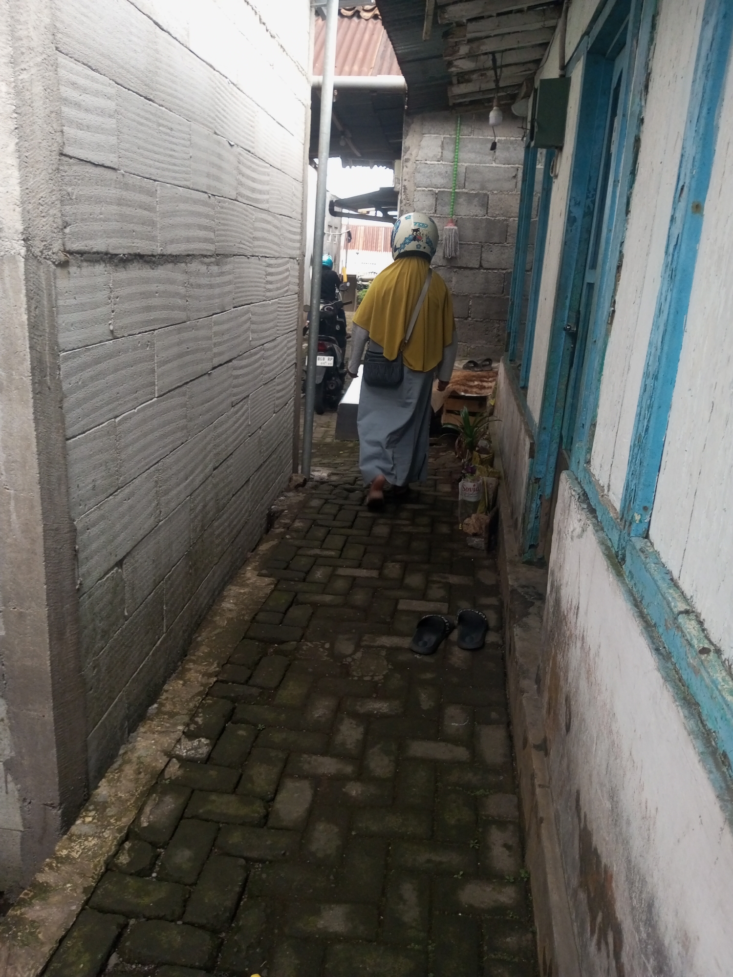 An overhead shot captures a narrow alleyway between two buildings in Andongsili, Indonesia. On the left, a textured concrete wall forms a stark barrier, its horizontal blocks marked with irregular indentations. To the right, a white wooden structure with blue trim lines the alley. A person wearing a yellow hijab and a grey skirt walks away from the camera, their back turned. They also wear a helmet with a cartoon decal and carry a grey bag slung over their shoulder. The ground is paved with dark bricks, some of which are covered in moss. A pair of black flip-flops rests on the brick pathway. In the background, a motorcycle is partially visible, suggesting a residential or urban setting.
