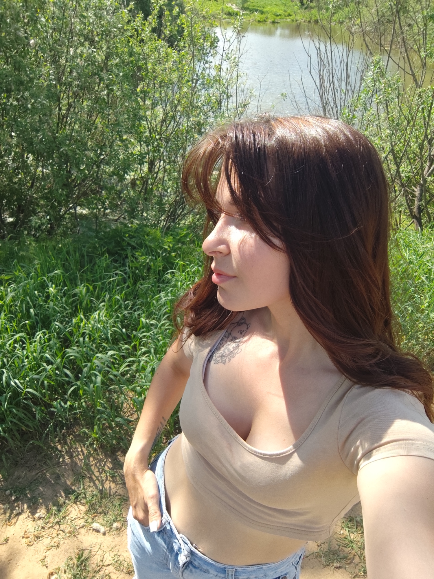A medium, eye-level shot depicts a woman standing outdoors near a body of water. The woman has long, brown hair with bangs, fair skin, and is seen in profile. She is wearing a beige, short-sleeved crop top and light-wash blue jeans, with one hand casually placed in her pocket. A tattoo is visible on her shoulder.
The background includes green vegetation such as bushes and tall grass. The body of water is a light green color, likely reflecting the sky and surrounding foliage. The ground appears to be dirt and dry leaves. The location is Фоки, Russia.