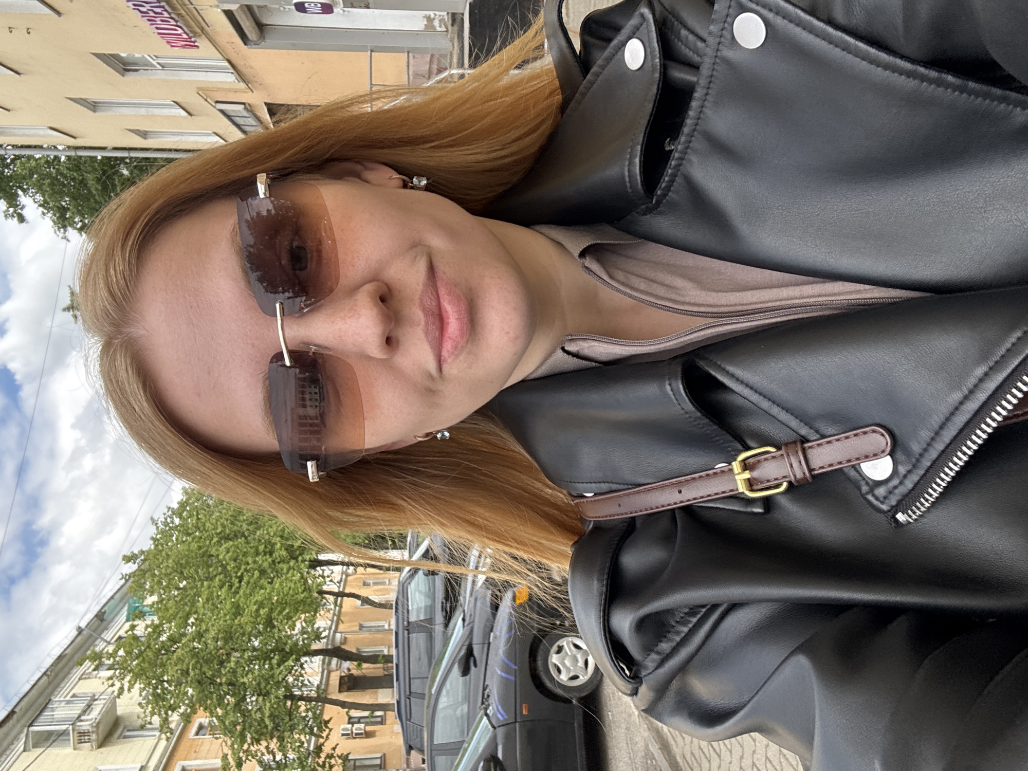 A woman with fair skin and long, light-brown hair is wearing sunglasses and a black leather jacket. She has on a light-colored top under the jacket. The photo appears to be a selfie taken outside, with buildings and trees visible in the background. A dark-colored vehicle is parked on the street. The photo was taken in Заволжье, Russia.