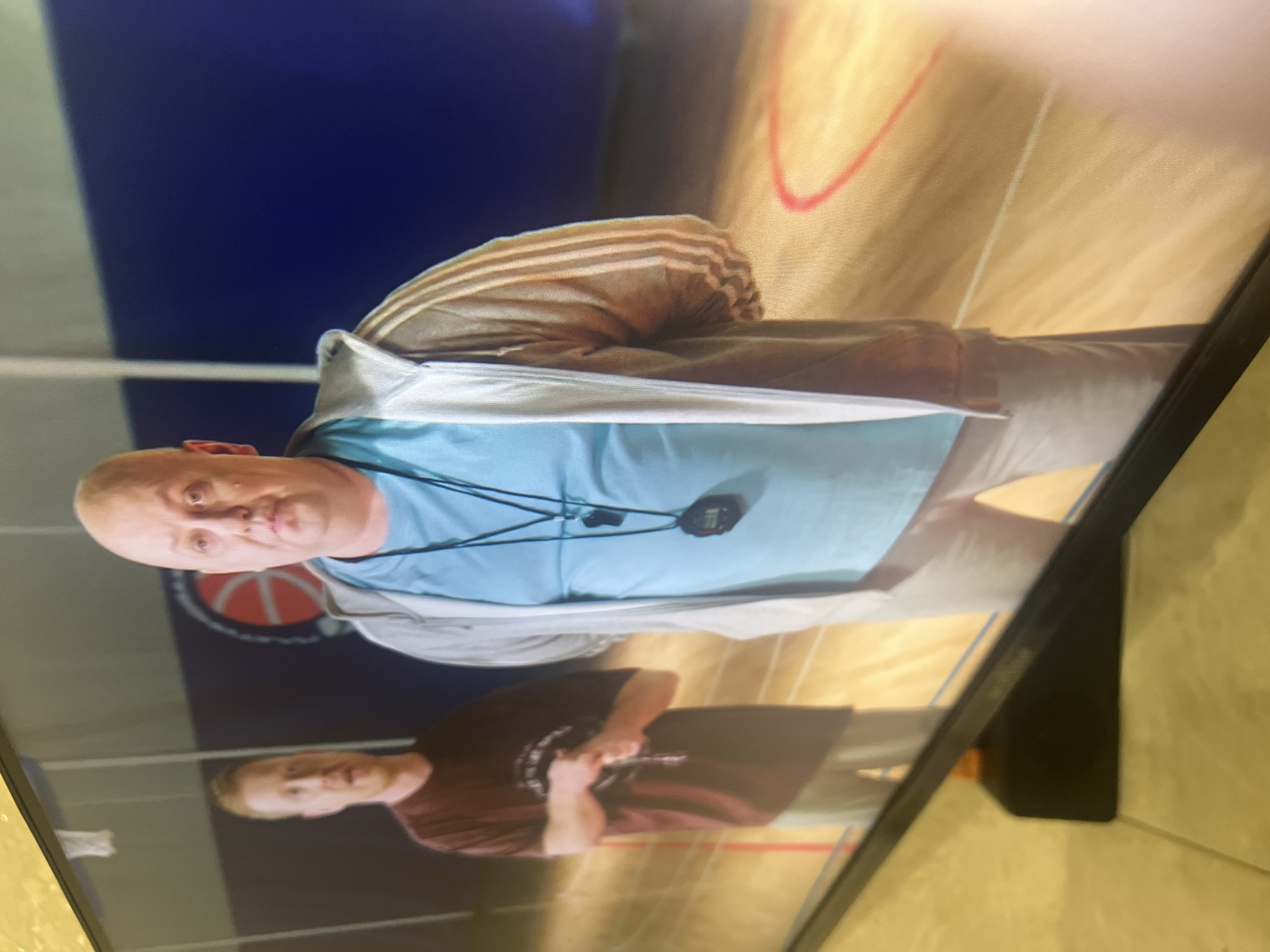 Two men are standing on a basketball court. The man on the right is bald, wearing a light blue t-shirt and a gray zip-up hoodie. He has a whistle on a lanyard around his neck. The man on the left is wearing a maroon t-shirt and light-colored pants. He is looking at his hands, which are clasped in front of him. The court has a wooden floor with red and white lines. In the background, there is a blue wall with a basketball hoop and a large circular logo with a basketball graphic. The scene appears to be indoors. There is no visible indication of the location being in Saratov, Russia.