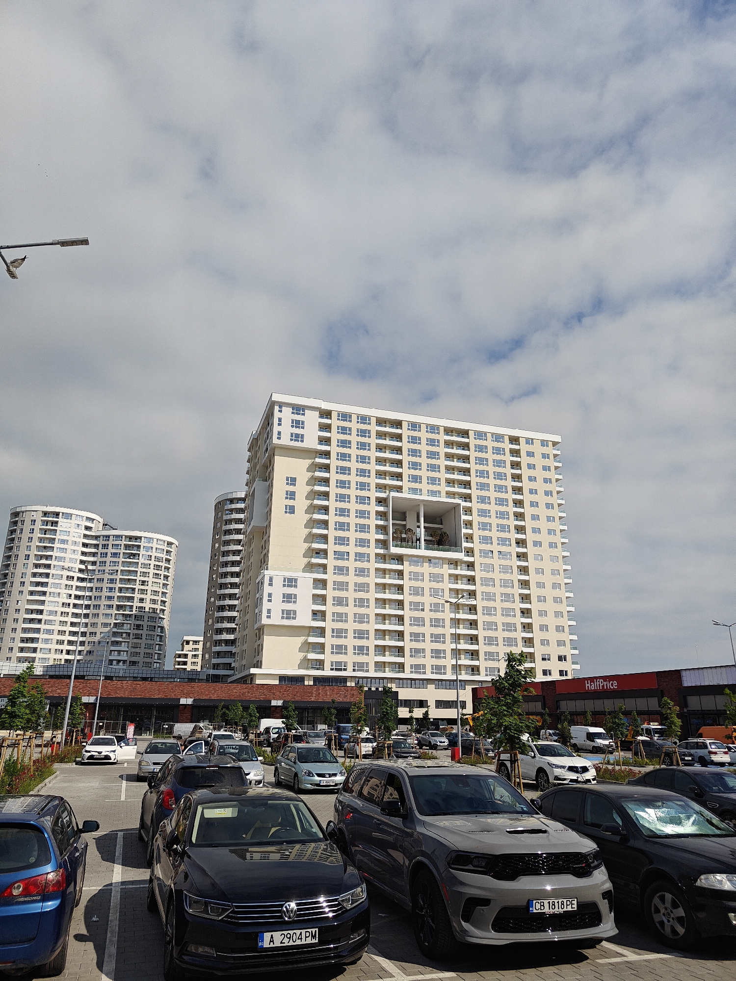 The image shows a parking lot in Burgas, Bulgaria, with several cars parked in marked spaces. In the background, there are several high-rise apartment buildings and a shopping center identified by a "HalfPrice" sign. The sky is cloudy with patches of blue. A lamp post is visible on the left side of the image. The focus is on the cars in the foreground, with clear visibility of license plates. The overall scene depicts a typical urban environment with commercial and residential elements.