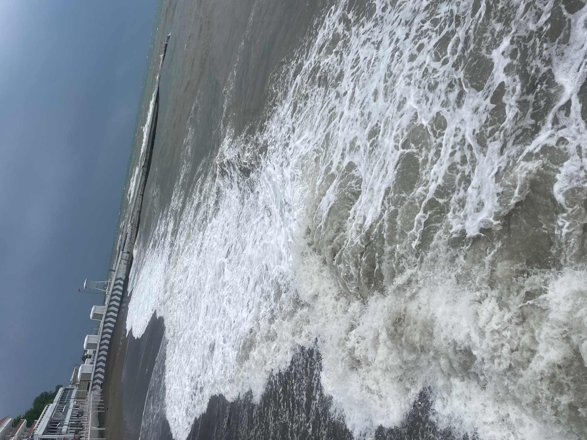 The image depicts a beach in Sochi, Russia, on a cloudy day. Waves are crashing onto the shore with white foam. A pier or breakwater extends into the sea, parallel to the beach. Beachfront buildings and a lifeguard tower are visible on the left. The sky is overcast.