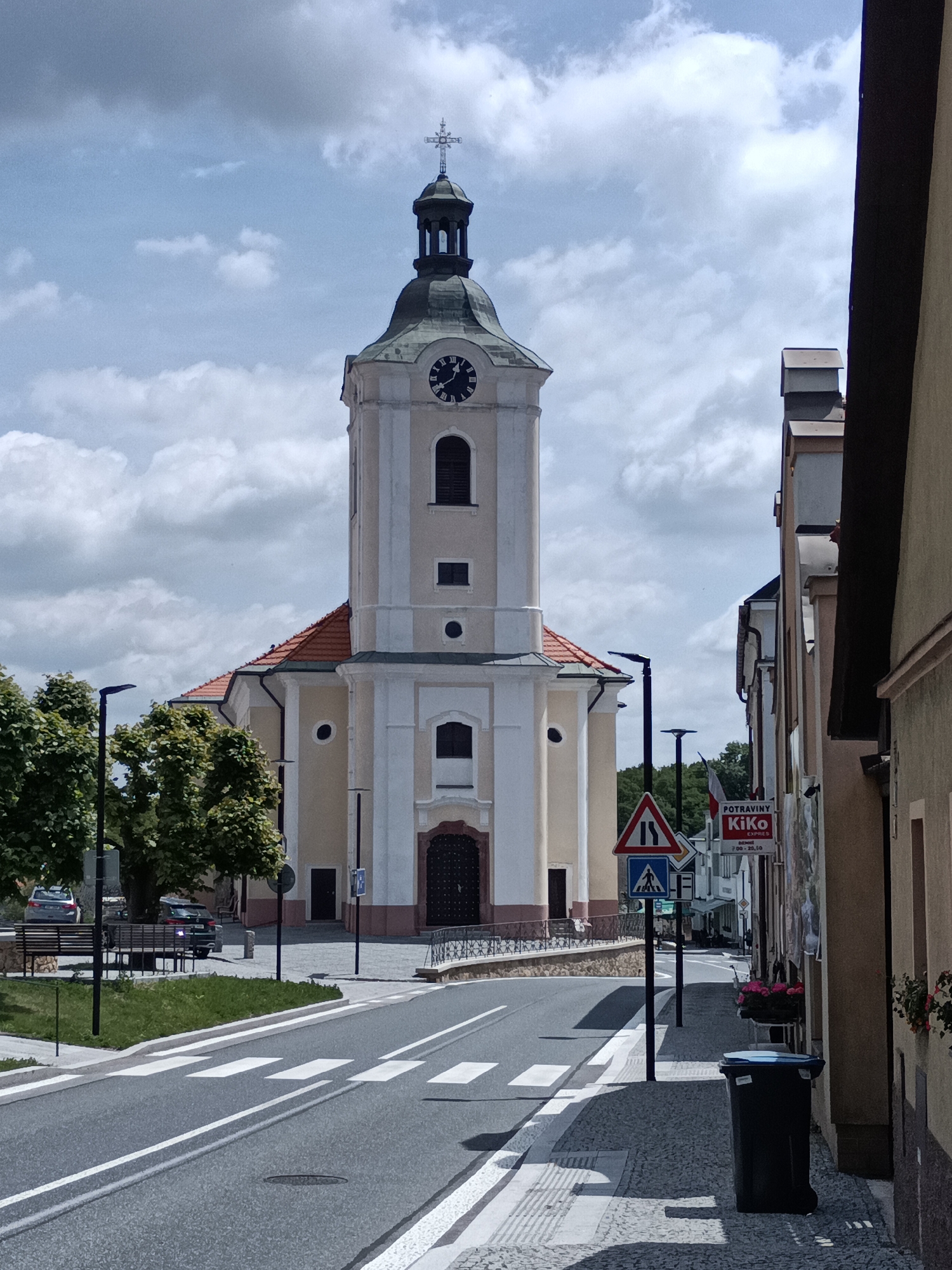 The image shows a street scene in Dalovy, Czechia, dominated by a large, pale yellow and white church with a tall clock tower. The church has a red-tiled roof and a dark arched doorway. The street in front of the church is paved with asphalt and marked with a pedestrian crossing. To the right of the street, there is a sidewalk made of cobblestones, with a black trash can and planters with flowers. Several traffic signs are visible, including a yield sign and a pedestrian crossing sign. Trees line the left side of the street, and a bench is situated on a patch of grass. A few cars are parked on the side of the road. The sky is partly cloudy.