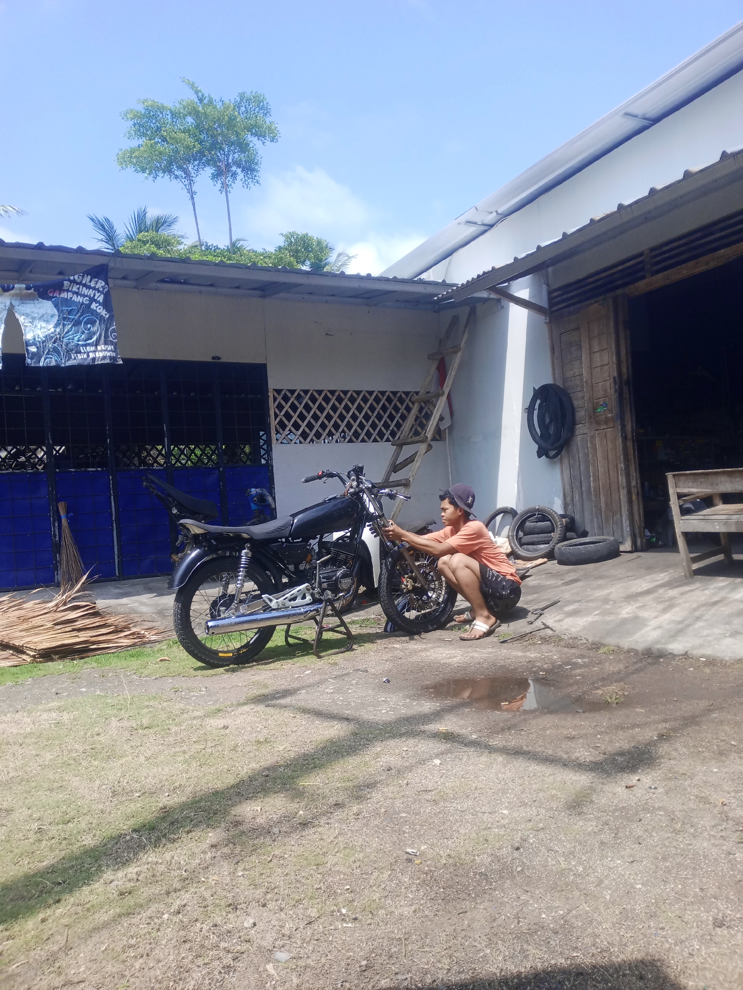 A young man, wearing an orange t-shirt, black shorts, a black cap, and white sandals, is crouched down working on a black motorcycle. The motorcycle is propped up on a stand. The man appears to be making adjustments to the front wheel or handlebars. Behind him, a ladder leans against the side of a white building with a corrugated metal roof. To the left of the building, there are blue metal gates. A pile of palm fronds or similar material is stacked next to the gates. To the right of the building, there is an open doorway, likely leading into a shop or garage, with wooden doors. Several tires are stacked near the doorway, and a wooden bench is visible inside. The ground is a mixture of dirt and grass, with a small puddle of water reflecting the sky and building. The scene suggests a rural or semi-rural setting, possibly a motorcycle repair shop, in Cintaratu, Indonesia. The sky is blue with scattered clouds.