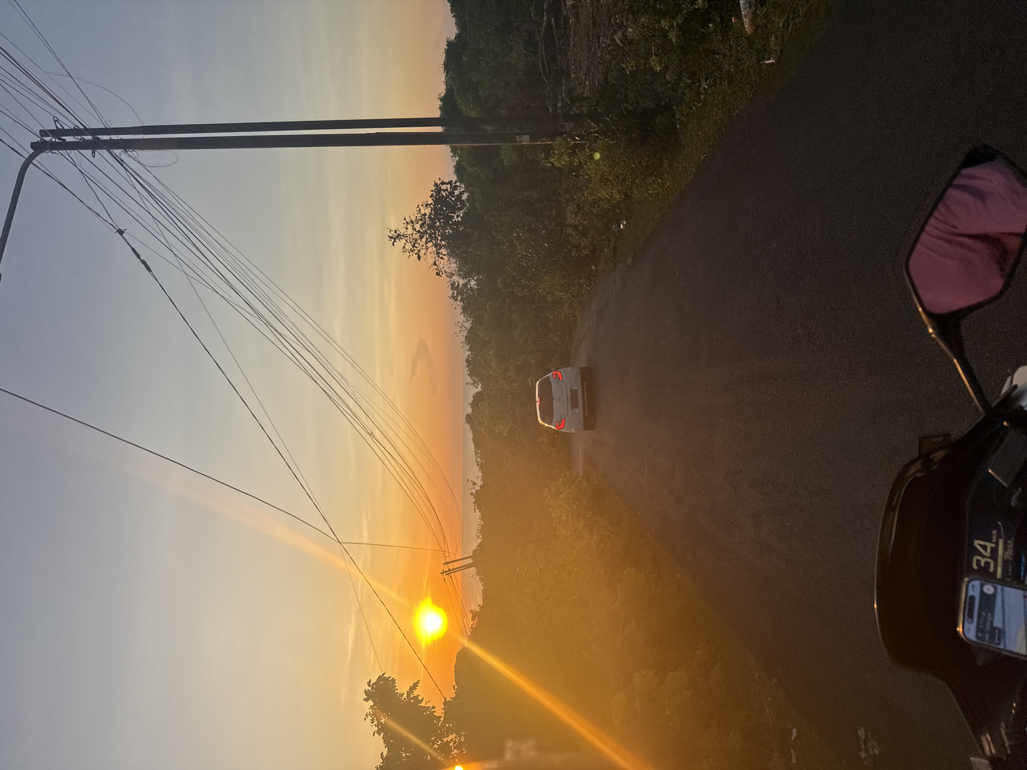 A photograph depicts a sunset viewed from a road, likely from a motorcycle. A white car is positioned on the road ahead. The sunset casts a warm orange glow across the sky and landscape. Power lines are visible on the left side of the frame. A side mirror of a motorcycle is visible on the right side of the frame. The dashboard of the motorcycle is also partially visible, displaying the number "34". The road is asphalt. The location is unknown.