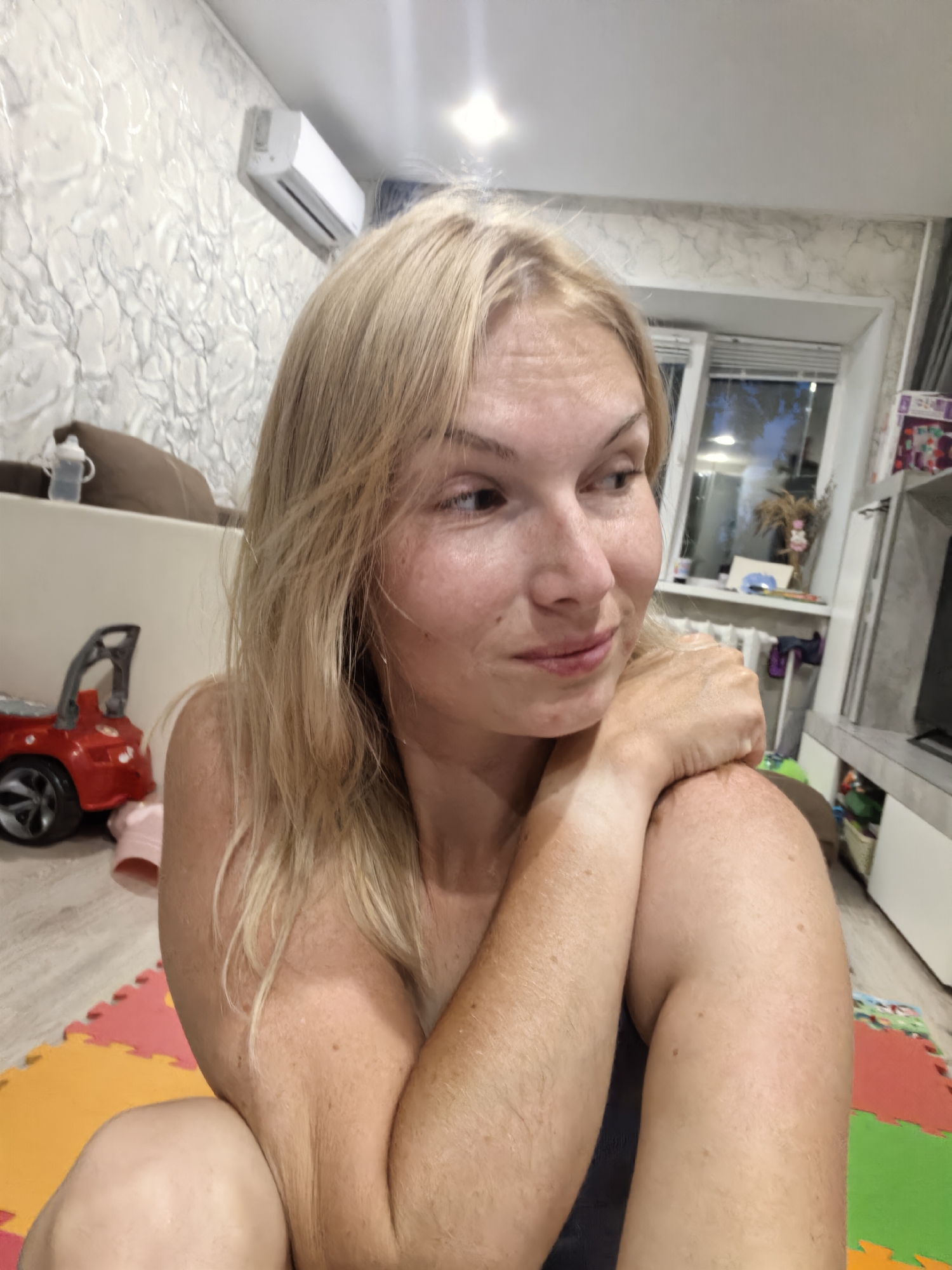 A medium shot captures a woman with blonde hair from a low angle. She is looking to her right with a slight smile. Her right arm is bent and resting on her shoulder, with her left arm visible on her side. The background shows a wall with textured wallpaper, an air conditioning unit, a window with blinds, and a toy car. A portion of a puzzle mat is visible on the floor in the foreground. The scene appears to be indoors, likely a residential setting. No information about the city of Sterlitamak, Russia, or its specific location within it is discernible from the image.