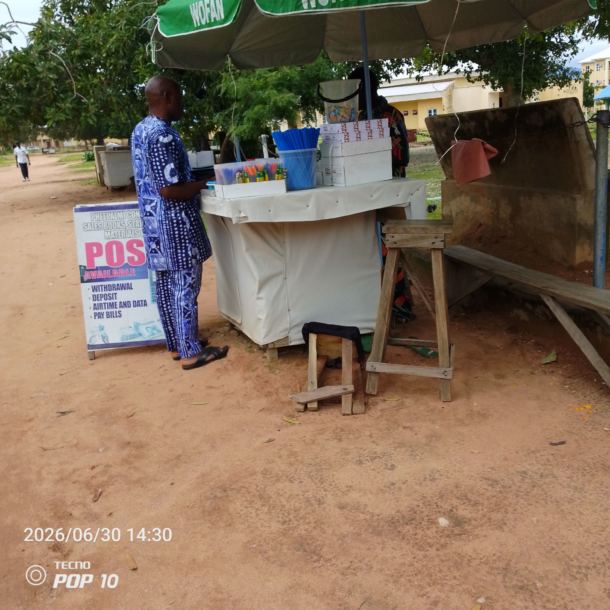 The image shows an outdoor scene in Jalingo, Nigeria, possibly a street or a market area. The focal point is a temporary stall set up under a green and white umbrella with "WOFAN" printed on it. A man in a blue and white patterned traditional outfit is standing behind the stall. The stall itself is covered with a white cloth and displays various items, including stacks of boxes with "POPC" and "POPO" visible, a bucket filled with what appears to be candy or small packaged goods, a blue container with blue straws, and smaller containers with pens and other small items. A sign in front of the stall advertises "POS AVAILABLE" with services like "WITHDRAWAL," "DEPOSIT," "AIRTIME AND DATA," and "PAY BILLS."
In the background, there are trees, and in the distance, some buildings, suggesting a semi-urban or institutional setting. Another person, wearing a white shirt, is walking away from the stall in the left background. The ground is sandy or dirt. The time stamp at the bottom of the image reads "2026/06/30 14:30" and a "TECNO POP 10" logo is also visible. The weather appears to be overcast or cloudy, given the lighting. The scene depicts a commercial activity, likely a point-of-sale service provider operating from a roadside stall.