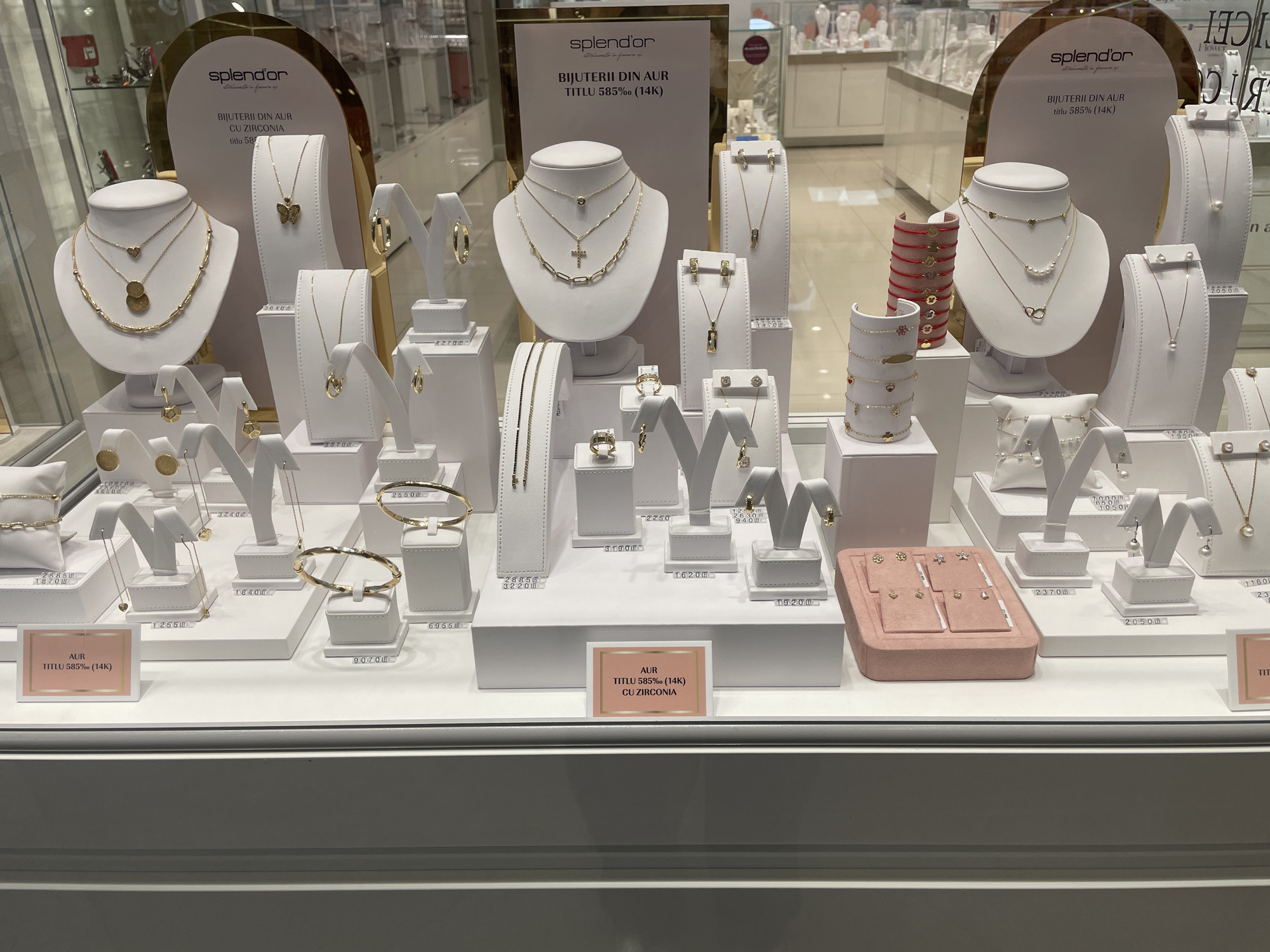 The image shows a well-lit display window of a jewelry store, likely located in Iași, Romania, as suggested by the product labels in Romanian. The display features a variety of gold jewelry, including necklaces, bracelets, earrings, and rings, presented on white mannequins and display stands. The jewelry is primarily gold and some pieces appear to have gemstones or cubic zirconia. The store's branding, "splendor," is visible on a prominent display board. The background shows other display cases and the interior of a store. There are no people visible in the image. The overall impression is one of a clean, organized, and upscale jewelry retail environment.