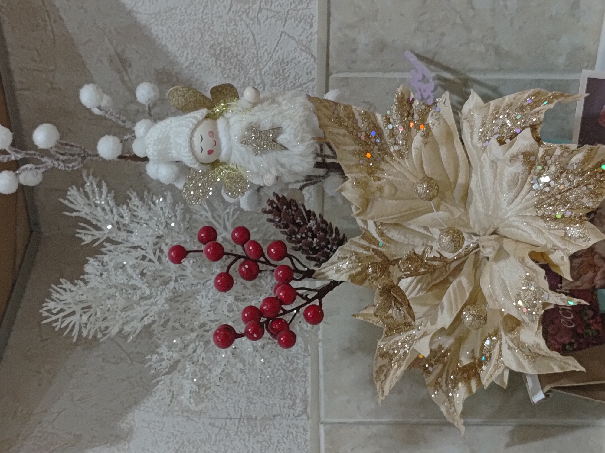 The image shows a festive, close-up shot of a decorative arrangement, likely for a holiday season. The focal point is a small angel figurine with a white knitted hat, golden glittery wings, and a white fluffy robe adorned with a golden star. The angel is positioned among various festive elements: a glittery white artificial tree branch, a cluster of red berries on a thin branch, and a small pinecone. Below these, a large, cream-colored artificial poinsettia with golden glitter accents dominates the lower part of the frame. The backdrop appears to be a tiled wall with a subtle textured pattern. The lighting suggests an indoor setting. There is no indication of the city of Saransk, Russia, or any specific location within it.