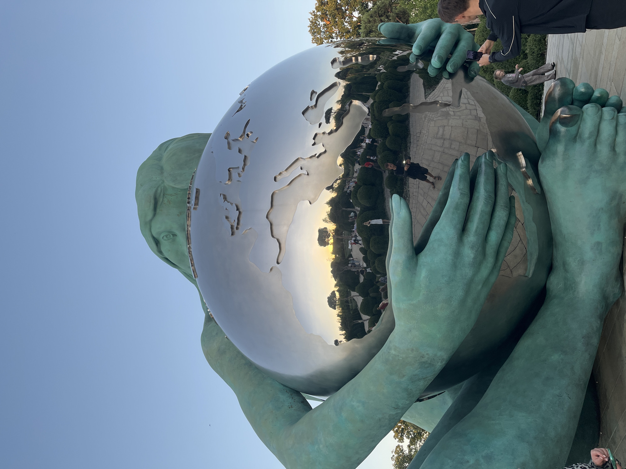 The image shows a close-up of a large, teal-colored statue of a giant figure cradling a reflective sphere that appears to be a map of the Earth. The statue is made of metal and has a weathered, greenish patina. The sphere reflects the surrounding environment, including a sunset, trees, and people. In the foreground, two hands of the statue are visible, holding the sphere. A man in a black jacket is standing to the right, taking a photo of the statue with his phone. A young girl in a grey tracksuit is visible further back, walking away from the camera. The background shows a park-like setting with manicured bushes and trees, and a clear blue sky with a hint of sunset colors. The statue is located in Krasnodar, Russia.