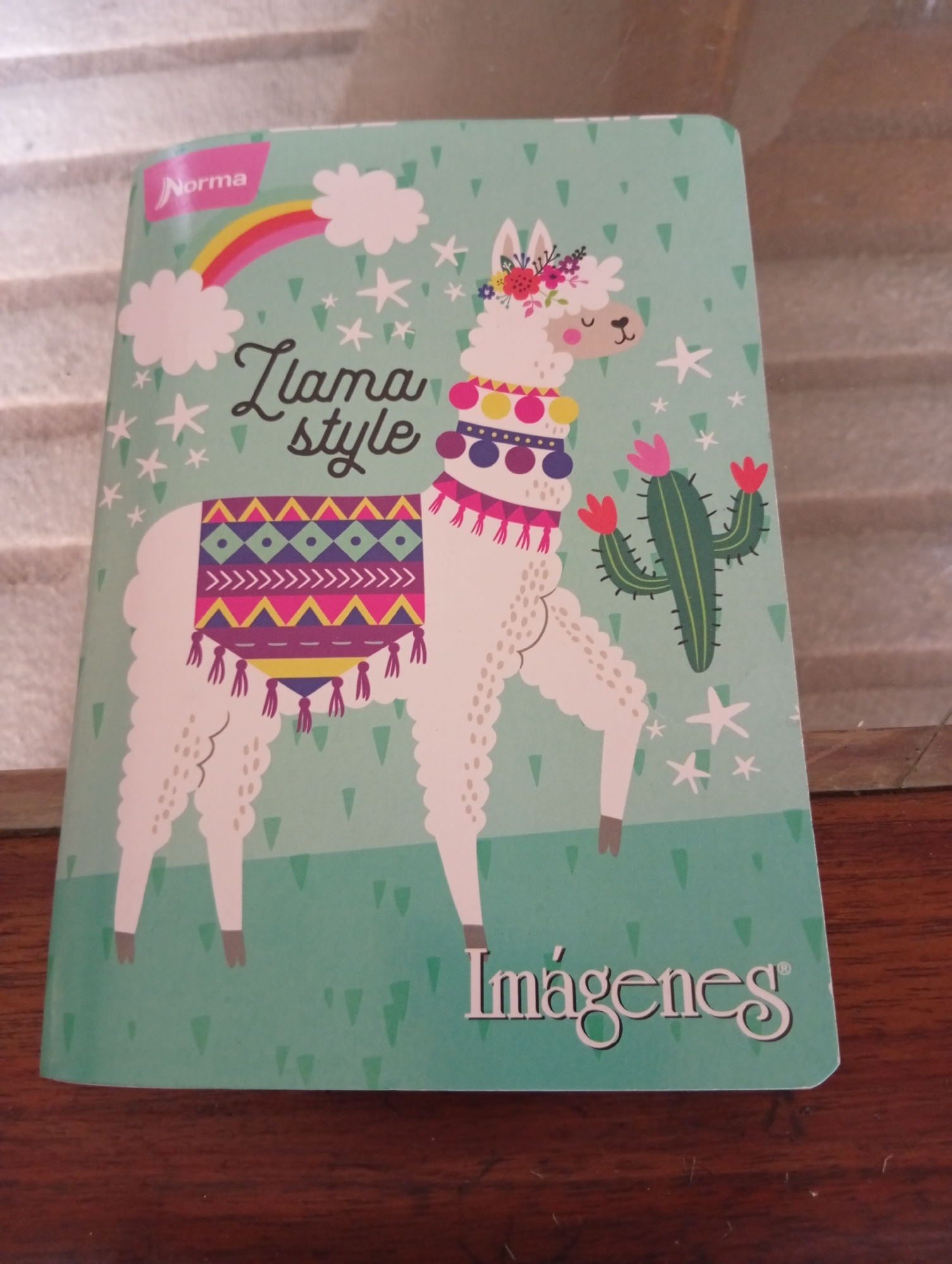 The image shows a close-up of a notebook with a teal background, featuring a cartoon illustration of a llama adorned with a colorful saddle and a floral headdress. Next to the llama is a cactus with pink flowers. The notebook has "Norma" printed on a pink banner in the upper left corner and "Llama style" written in cursive text. The bottom right corner of the notebook displays the word "Imágenes" in white stylized font, followed by a registered trademark symbol. The notebook is placed on a wooden surface, possibly a table, and the background behind it appears to be a textured surface with horizontal lines. The scene does not provide any specific information or context that would allow for its location to be identified as San Antonio de Los Altos, Venezuela.