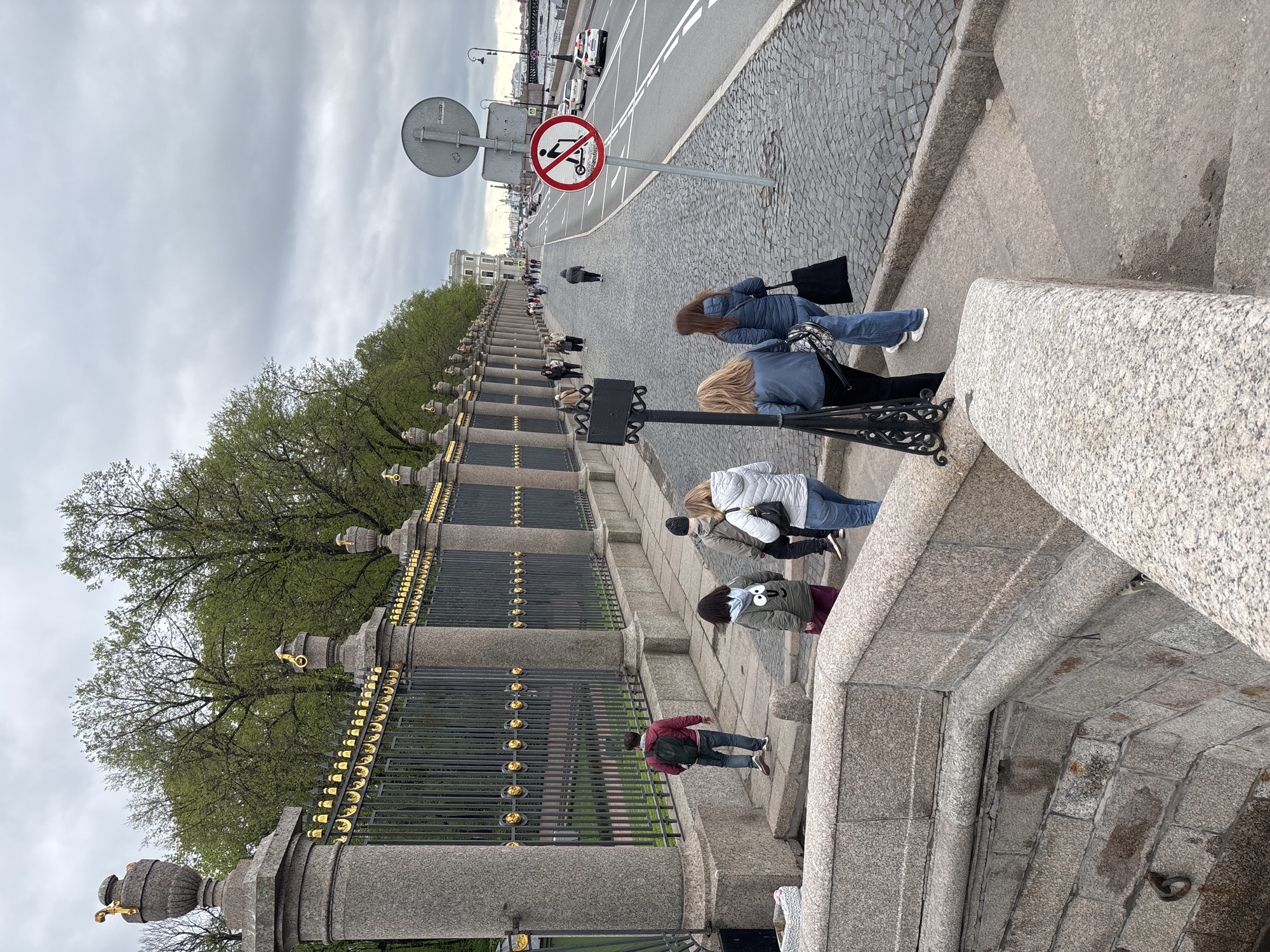 The image captures a scene on a street in Saint Petersburg, Russia. The focus is on a wide set of stone steps adjacent to a road. A decorative iron fence runs along the side of the steps, adorned with gold accents. Several people are walking along the steps.
On the right side of the image, a road sign is visible, indicating "No electric scooters." The road itself is paved with cobblestones. The sky is overcast, suggesting a cloudy day. In the background, buildings can be seen lining the street.