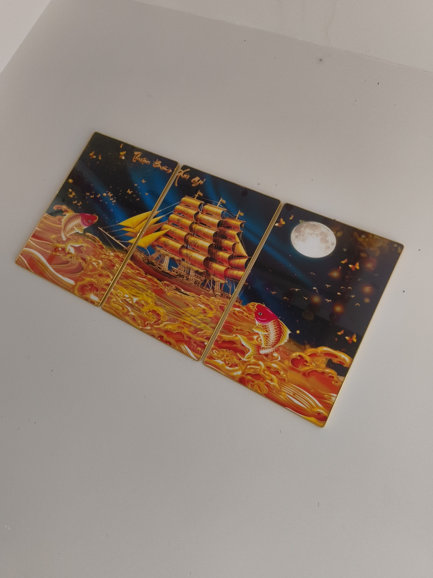 The image shows a triptych artwork featuring a sailing ship with yellow sails on a dark blue sea. The artwork is divided into three panels. The left panel depicts a red and orange fish jumping out of golden waves. The middle panel shows the sailing ship. The right panel features a large full moon, a flock of birds, and butterflies against a dark sky, with another red and orange fish in golden waves at the bottom. The artwork is placed on a light-colored, flat surface. The image is taken from a slightly elevated angle, looking down at the artwork. There are no people or animals present in the scene besides the depicted subjects in the artwork. The environment appears to be indoors with plain walls. The artwork is likely a decorative piece intended for display in a home or establishment in Phú Quốc, Vietnam, given the presence of Vietnamese script on the artwork itself.