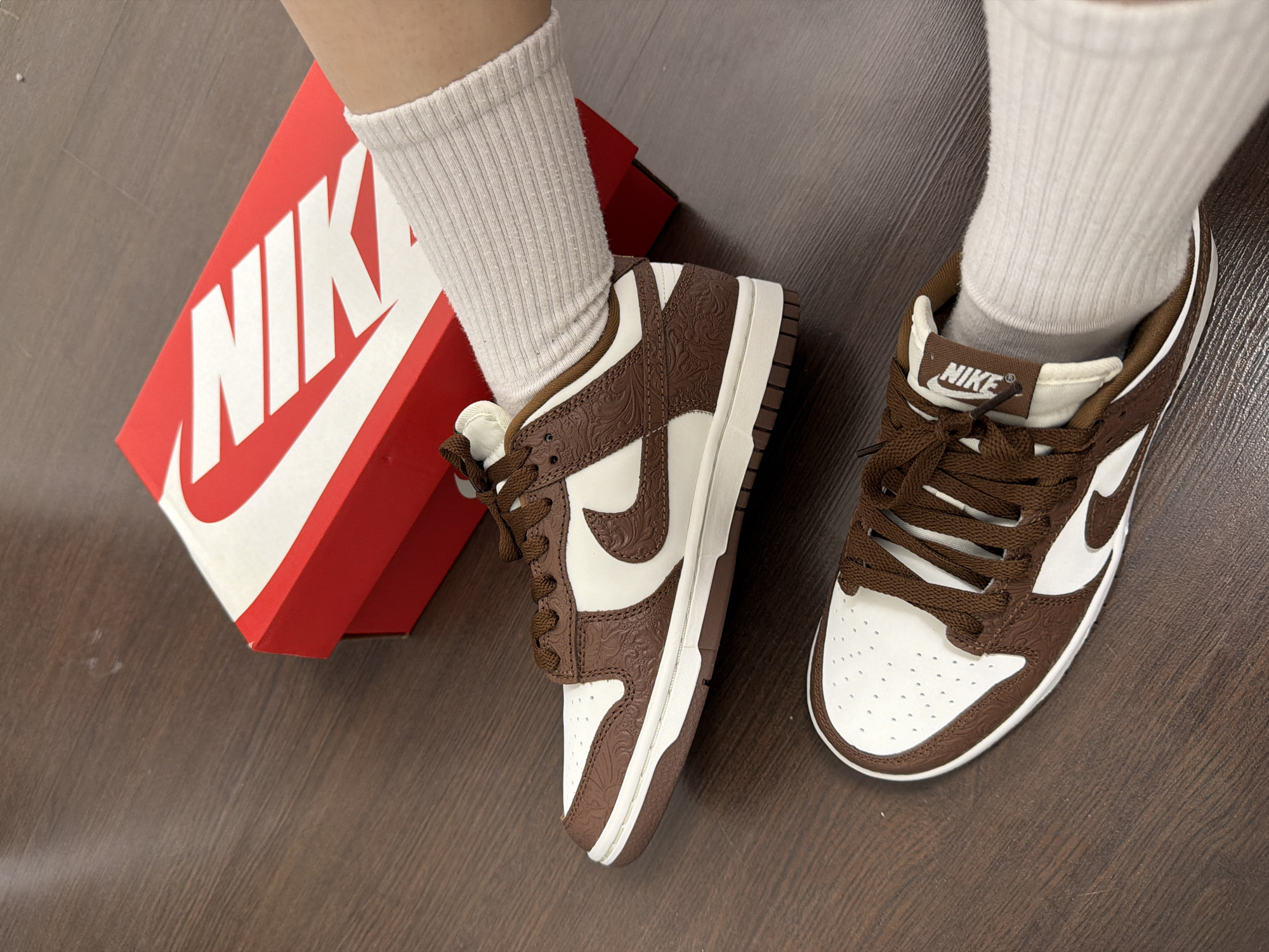 The image shows a pair of Nike Dunk Low sneakers in a brown and white color scheme, with a floral pattern on the brown leather overlays. The shoes are worn with beige ribbed socks. A red and white Nike shoebox is visible beside the left shoe. The background appears to be a wooden floor. There is no evidence to suggest that the photo was taken in Yekaterinburg, Russia.