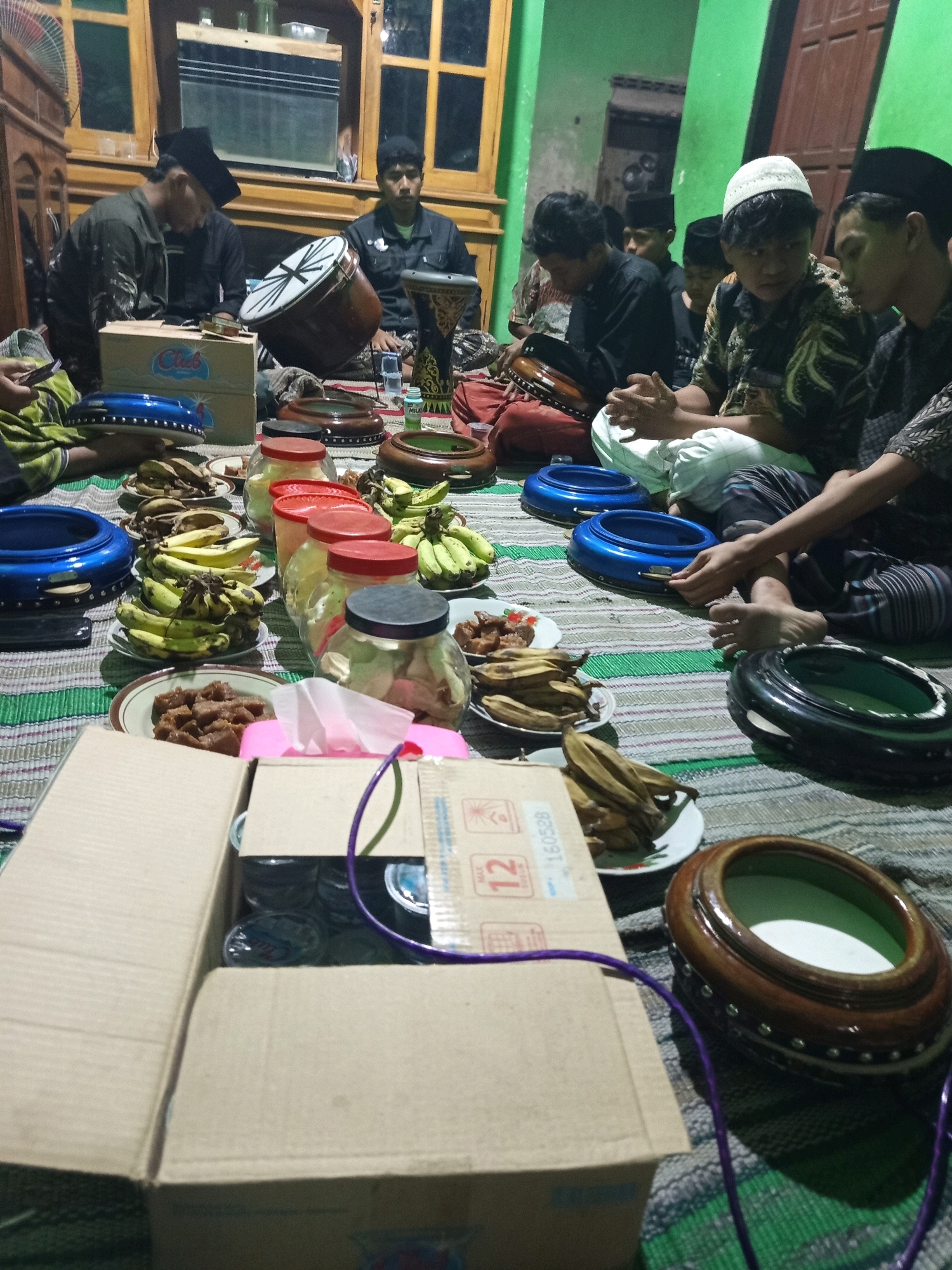 In a dimly lit room, likely in Mrican, Indonesia, a group of young men are gathered on the floor, engaged in what appears to be a cultural or religious activity. They are seated on a woven mat, surrounded by various musical instruments, primarily drums of different sizes and styles, including what look like frame drums and goblet drums. Several individuals are holding or interacting with these drums, suggesting they are either playing or preparing to play them.
The scene is also adorned with food items. Plates of bananas and what seem to be dried okra are visible, along with several jars containing food and some red-lidded containers. A cardboard box, some of which is open to reveal contents, and a box labeled "Club" are also present in the foreground, suggesting a communal gathering or event. The room's ambiance is further enhanced by wooden furniture, including what looks like a cabinet and window frames, contributing to a traditional Indonesian aesthetic. The lighting is dim, with some light coming from an overhead source and possibly from the windows, casting shadows and creating a somewhat intimate atmosphere. The participants are dressed in traditional attire, including head coverings like peci, adding to the cultural context of the scene. The overall impression is one of community and shared cultural practice within a domestic setting.