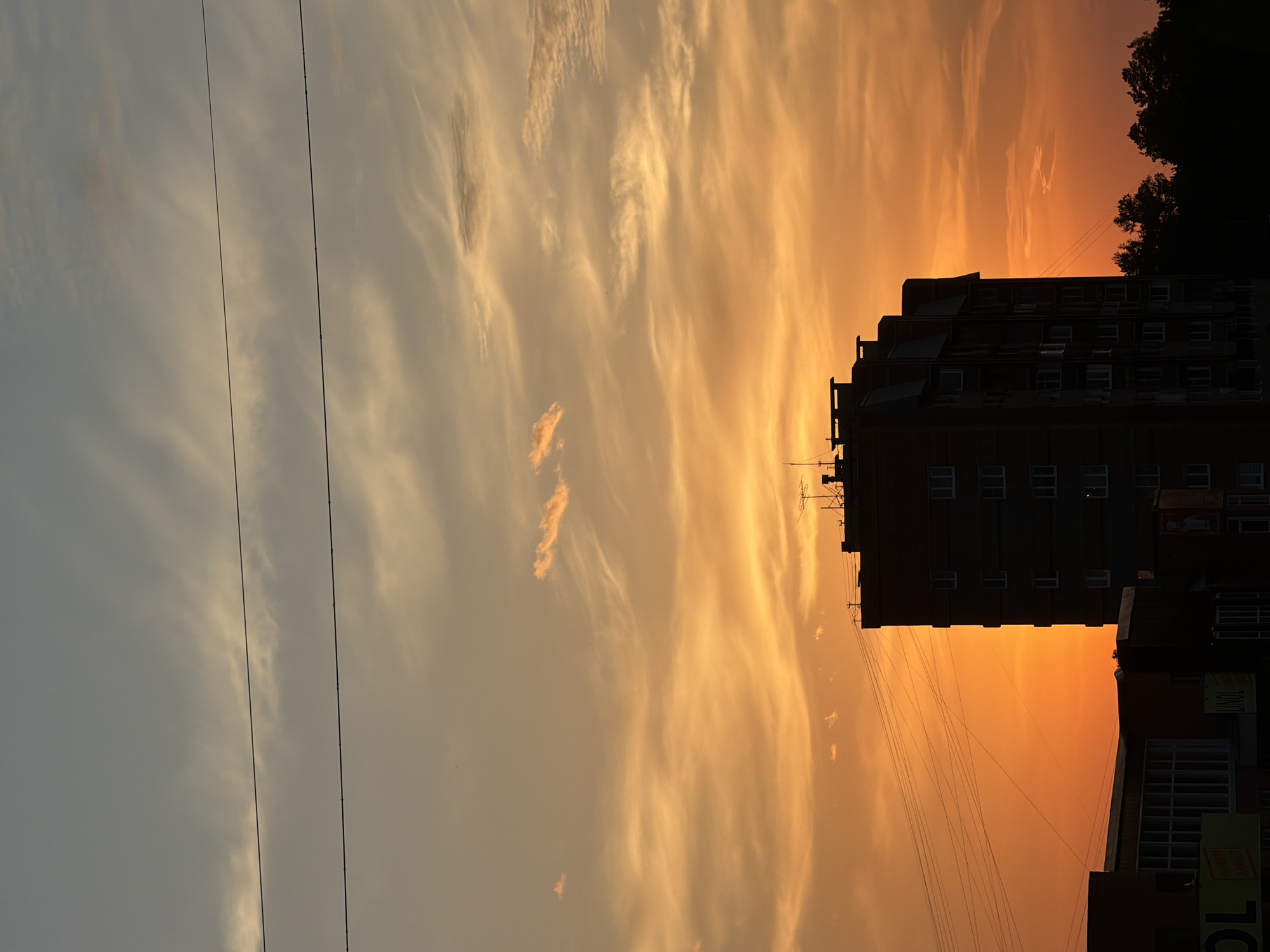 A vertical, medium shot captures the silhouette of a tall, brick apartment building against a dramatic sunset sky. The sky transitions from a light blue at the top to a vibrant orange and yellow closer to the horizon, with wispy clouds scattered throughout. Several thin, dark power lines stretch horizontally across the frame. In the foreground, the dark outlines of lower buildings and trees are visible, also silhouetted by the sunset. The apartment building, which appears to be in the city of Ust-Ilimsk, Russia, has a distinct architectural style with a pitched roof and numerous windows. No people are visible in the image. The overall atmosphere is serene and highlights the beauty of the natural light at dusk.