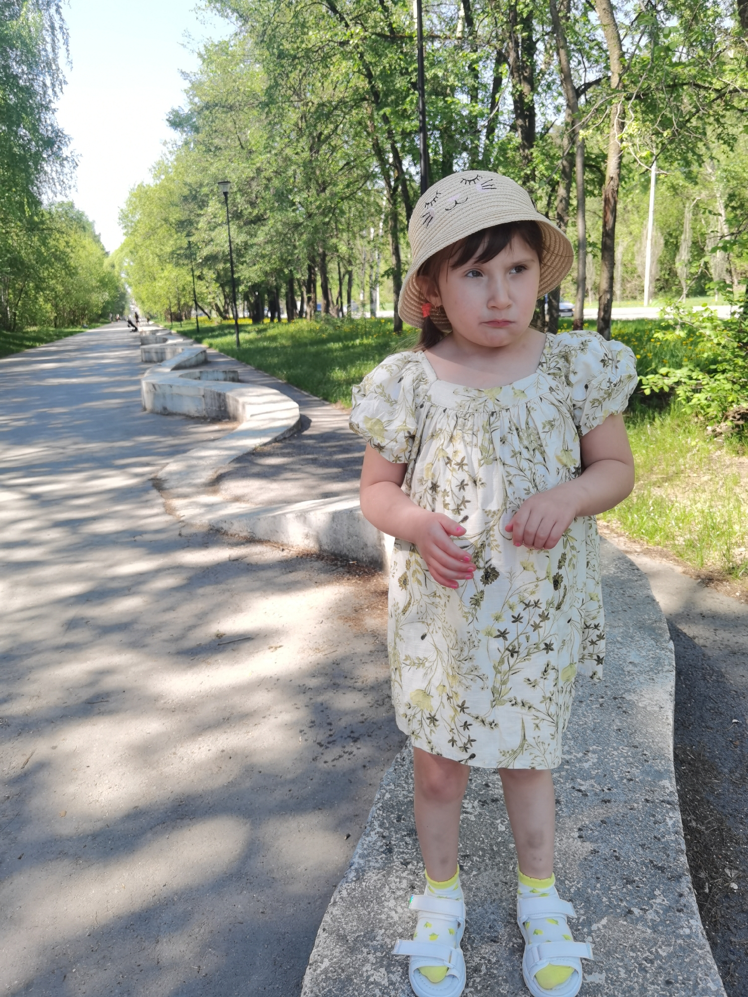 A young girl stands on a low concrete barrier at the edge of a paved path in a park. She is wearing a light-colored floral-patterned dress, white sandals with yellow socks, and a straw hat with a cat face design. The path stretches into the distance, lined with trees and concrete barriers along the edge. The location is in the city of Perm, Russia.