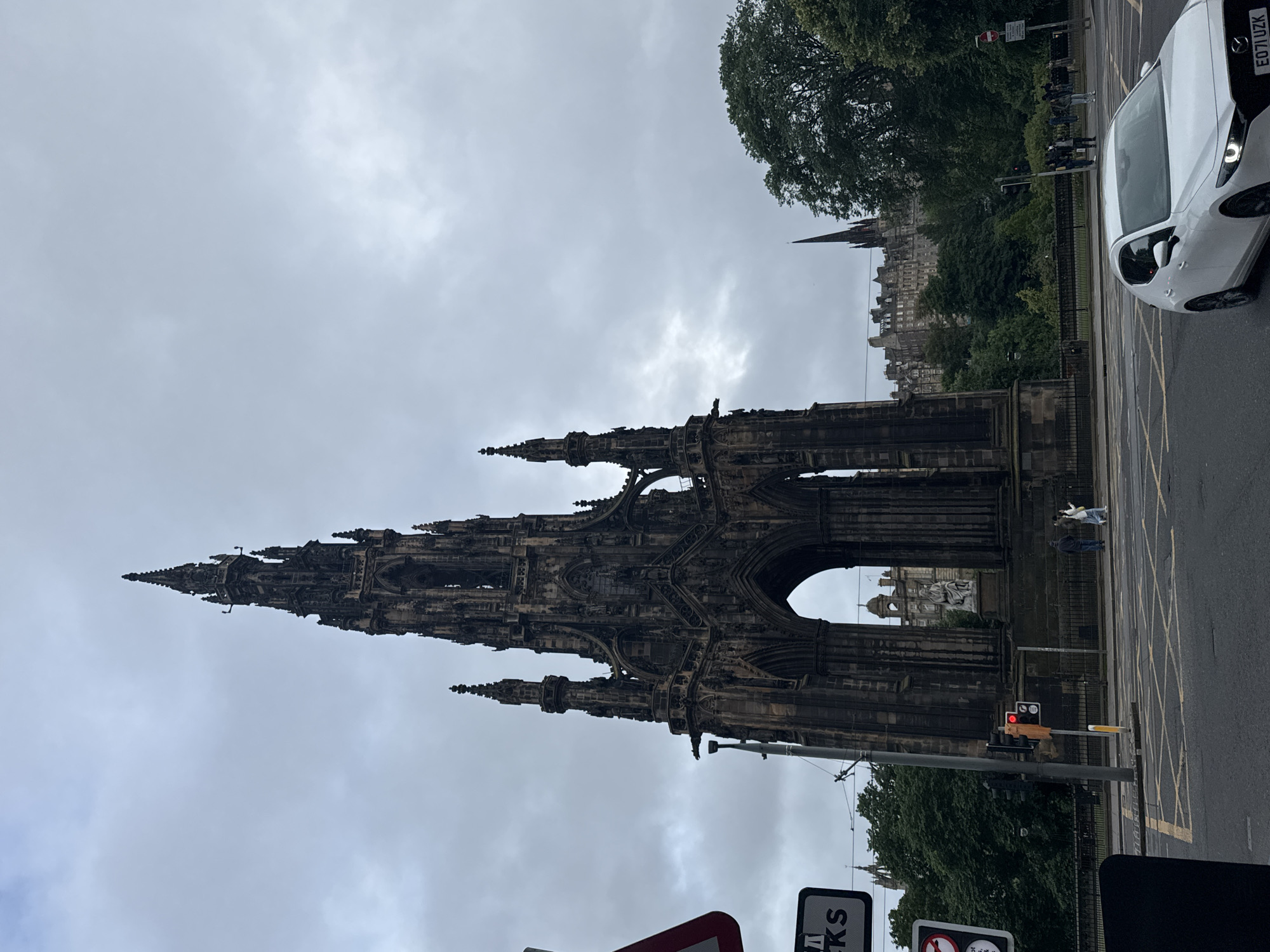 The image shows the Scott Monument, a Victorian Gothic monument to Scottish author Sir Walter Scott, located in Edinburgh, Scotland. The monument is a prominent spire constructed from red sandstone, with intricate carvings and a large archway at its base. In the foreground, a white car is driving on a wet road, indicating recent rain. Traffic lights and road markings are visible, suggesting an urban intersection. Several people are walking on the pavement and crossing the street. In the background, trees and buildings, including what appears to be the Edinburgh skyline, are visible under a cloudy sky. The overall atmosphere is overcast and somber.