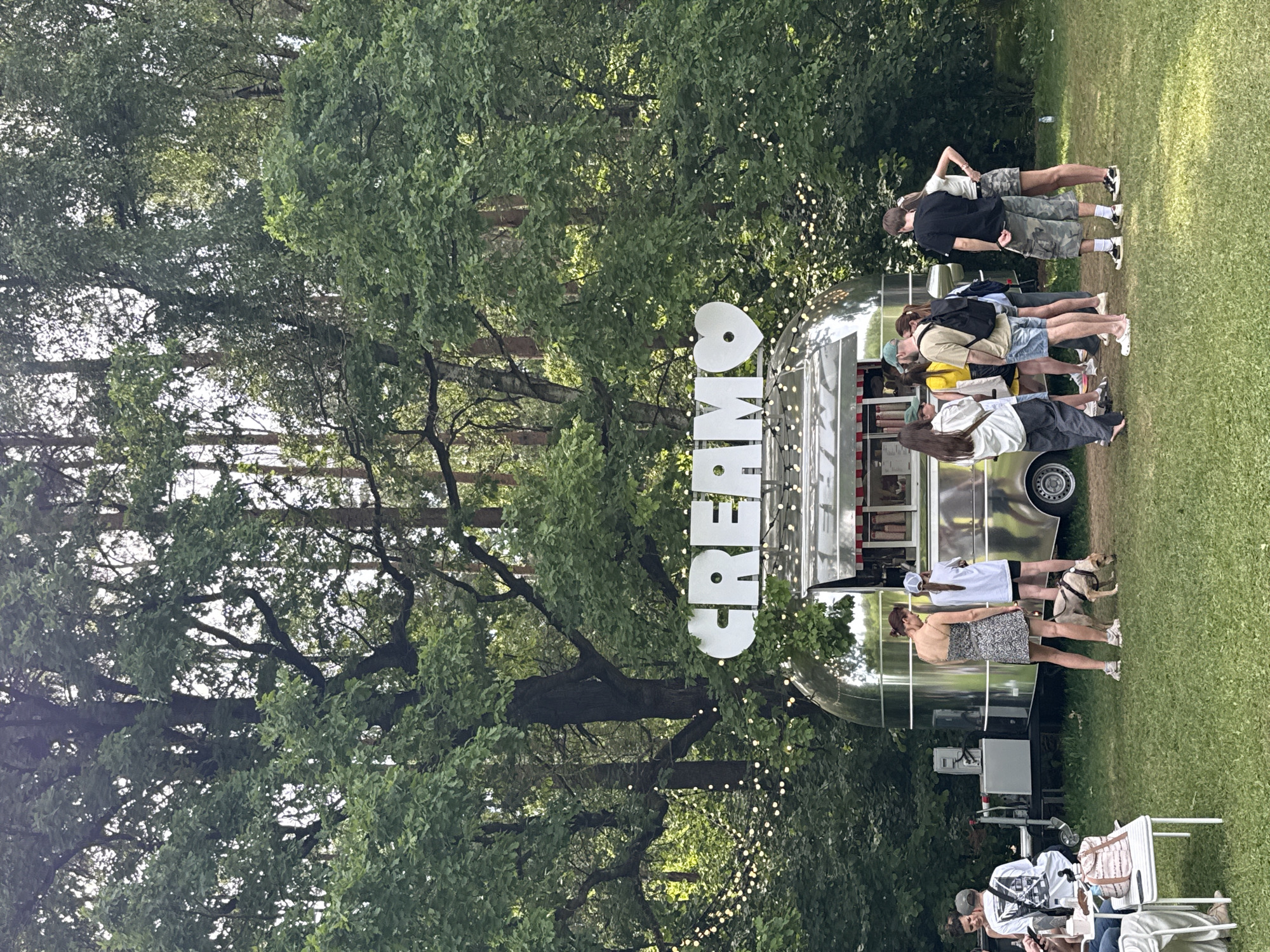 The scene depicts an outdoor food concession area in Nemchinovo, Russia, situated within a wooded park or forest environment. A polished silver, Airstream-style food trailer serves as the central focal point, featuring a large serving window. Above this window, white block letters spell "CREAM" followed by a white heart symbol. A string of small, warm white globe lights is draped across the front of the trailer and extends into the surrounding trees.
Approximately eleven individuals are present. A group of six to seven adults stands near the trailer's serving window. This group includes a woman in a patterned dress with a small, light-colored dog on a leash, which is sniffing the ground. A man wearing a green cap, a light-colored shirt, blue shorts, and a black backpack stands directly in front of the serving aperture. To his right, a man in a black t-shirt and camouflage shorts stands with his back partially to the trailer, alongside a woman in a white top and camouflage skirt. On the far left, two individuals are seated at a white rectangular outdoor table; one wears a white graphic t-shirt and a grey cap. A pink tote bag rests on this table. Adjacent to the left side of the food trailer, utility equipment, possibly a generator or power box, is visible on the grass.
The ground is covered with green grass, showing some wear in high-traffic areas around the trailer. Dense foliage from mature trees, including broadleaf and coniferous varieties, forms the backdrop, creating a shaded canopy. The scene is illuminated by natural daylight filtering through the trees.