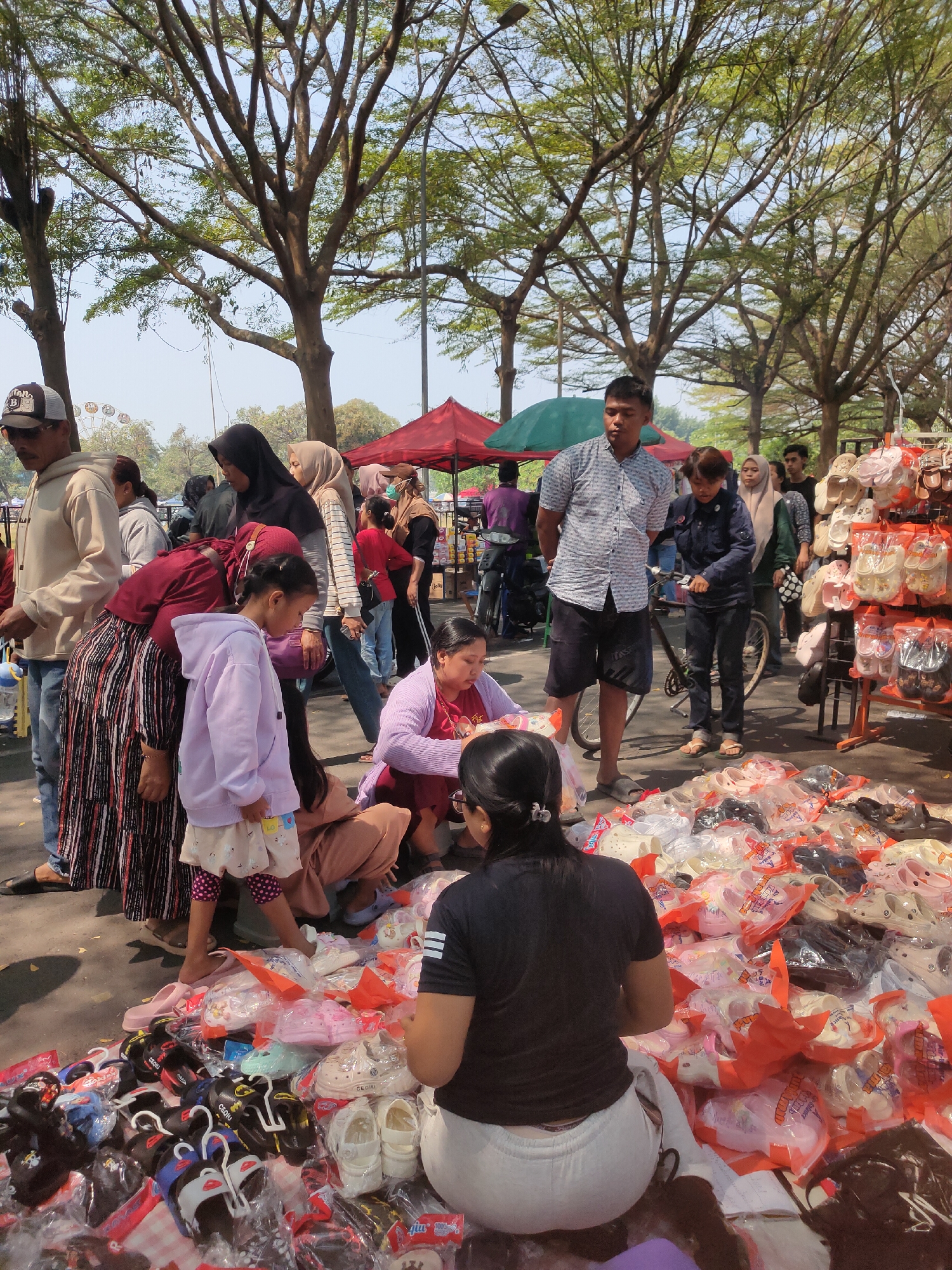 An outdoor market scene unfolds in the city of Ngasem, Indonesia, under the dappled sunlight filtering through the branches of large, mature trees. A diverse group of individuals, some wearing hijabs, are engaged in various activities, suggesting a bustling marketplace. In the foreground, a large pile of footwear, primarily sandals and clogs, is displayed in clear plastic packaging, interspersed with some loose items. A woman in a black t-shirt and white pants is seated, appearing to be tending to or organizing the merchandise. Nearby, a young girl in a light purple hoodie and patterned leggings stands, her gaze directed downwards. To the left, a man in a tan hoodie and a baseball cap is visible, partially obscured by other people. Further back, several individuals are browsing or walking through the market, with tents and stalls indicating commercial activity. A bicycle leans against a stall on the right side of the image, and in the background, more people and market structures can be seen, contributing to the lively atmosphere.