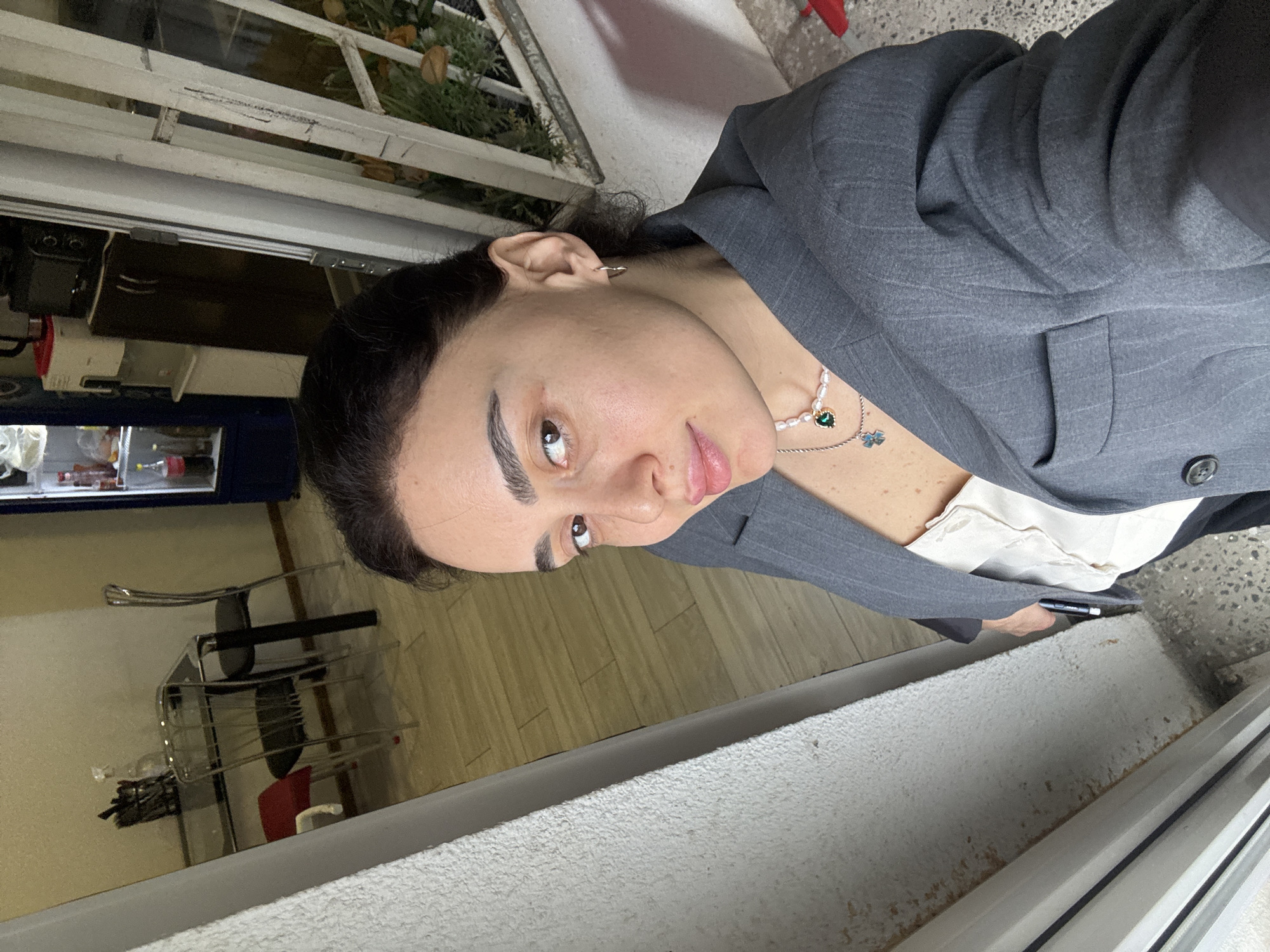 A woman in a gray blazer with a button is looking at the camera. She is wearing a pearl necklace with a green gem. The photo appears to be taken from a high angle. Behind her, a white railing with green plants is visible. To the left, there are glimpses of a room interior with wooden floors, a clear glass desk, and various objects on shelves. The location is possibly Tbilisi, Georgia, inferred from details such as architecture and vegetation typical of the region. She holds a pen in her right hand.