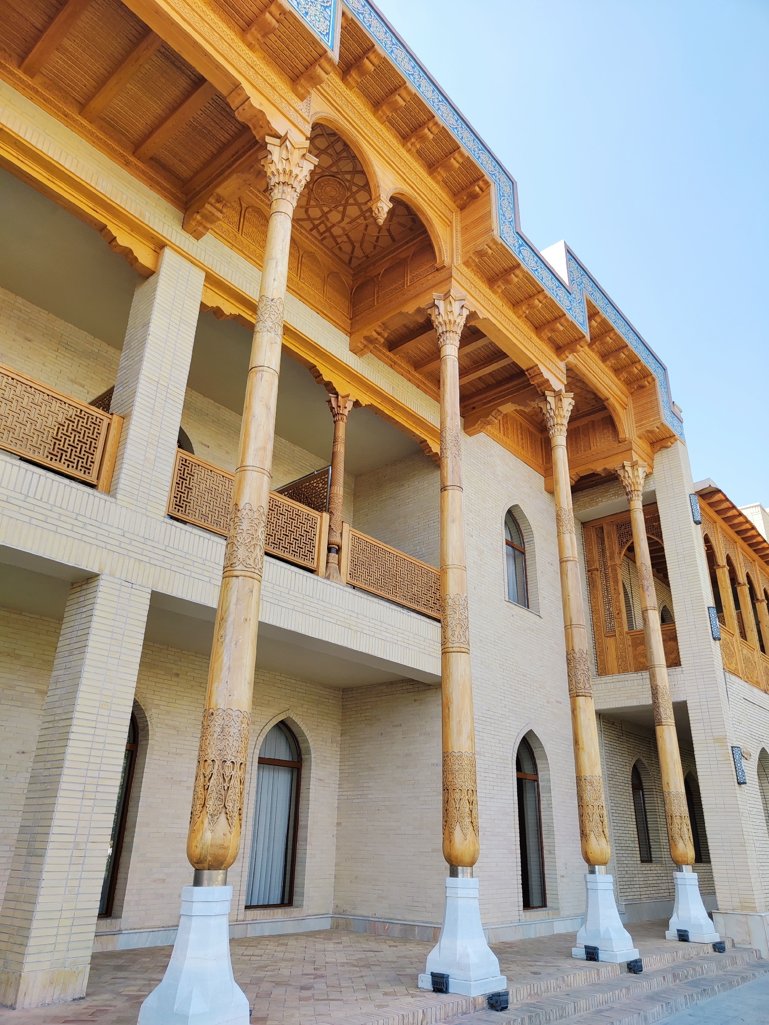 The image shows the exterior of a building in Tashkent, Uzbekistan, characterized by intricate wooden architectural elements. The building has a light-colored brick facade and is adorned with a wooden colonnade supporting an ornate wooden roof. The columns are thick and feature detailed carvings, resting on white bases. Wooden balconies with decorative screens are visible on the upper levels. Arched windows with dark frames are interspersed along the facade. The sky is clear and blue, indicating a daytime shot. The ground in front of the building is paved with bricks laid in a pattern. The overall impression is one of traditional Uzbek architecture with a focus on craftsmanship.