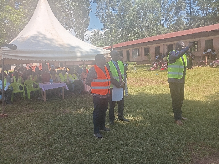 An outdoor gathering is depicted in the city of Idakho South ward, Kenya, on a sunny day. The scene unfolds on a well-maintained green lawn. On the left, a large white pyramidal canopy tent with scalloped edges covers an audience. Approximately 20-30 individuals are seated under the tent on light green plastic chairs; many are wearing yellow or light green reflective vests. A small wooden table draped with a light-colored tablecloth is positioned in front of some seated attendees.
To the right of the tent, three men stand in the foreground. The leftmost man, partially obscured, wears an orange reflective vest. The middle man wears a dark suit with a green reflective vest and holds papers. The rightmost man also wears a dark suit with a green reflective vest, holds a microphone in his right hand, and gestures with his left, appearing to address the assembled group.
In the background, a long, single-story building with a red corrugated iron roof and multiple windows is visible. A smaller group of people is gathered on the ground outside this building. Tall trees with green foliage and a bright blue sky with some haze complete the background.